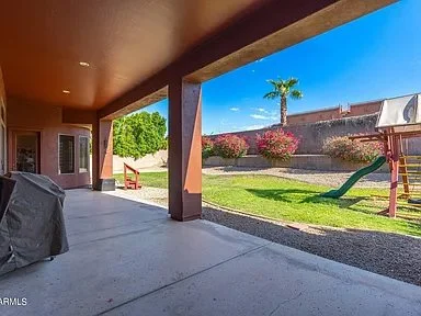Covered patio with a view of a backyard that has a palm tree, flowering bushes, a swing set, and a grassy area, under a clear blue sky.