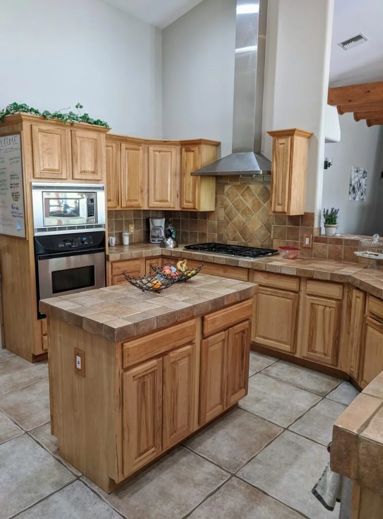 Kitchen with wooden cabinets, tiled countertops, a built-in microwave and oven, a stove with a range hood, and a small island in the center. There are potted plants and decorative items on the counters.
