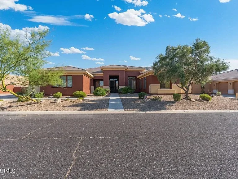 Single-story house with a desert landscape, two trees, and a driveway, under a blue sky with clouds.