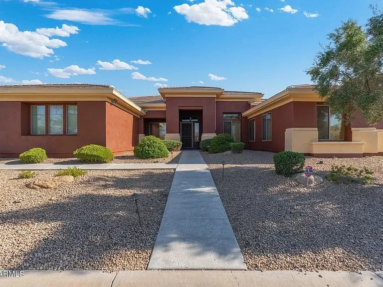 Front view of a single-story house with a concrete walkway leading to the entrance, surrounded by desert landscaping, bushes, and a tree, under a blue sky with scattered clouds.