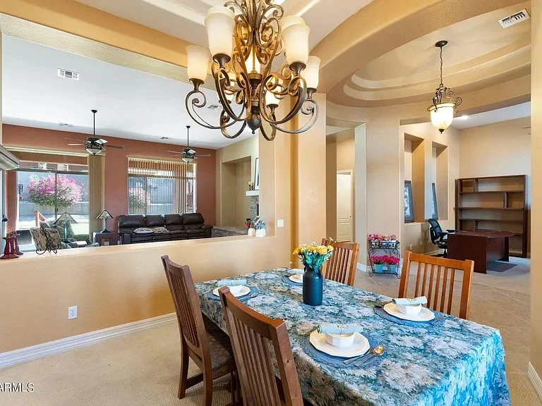 Dining area with a wooden table set with white plates and a blue vase filled with yellow flowers, surrounded by five wooden chairs, with a warm-colored interior and chandeliers hanging from the ceiling.