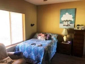 A bedroom with a bed covered in a blue quilt, a nightstand with a lamp and books, a window with blinds, and a painting of a tree on the wall.