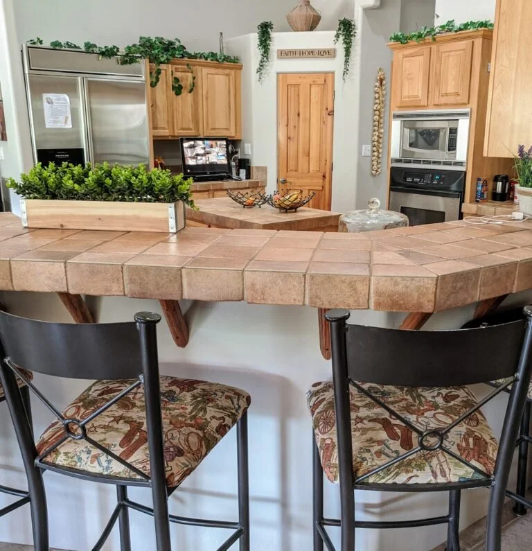 Kitchen with a curved tiled counter, two black chairs with patterned cushions, wooden cabinets, a stainless steel refrigerator, microwave, and various decorations including green plants and a sign that says 'Faith Hope Love'.