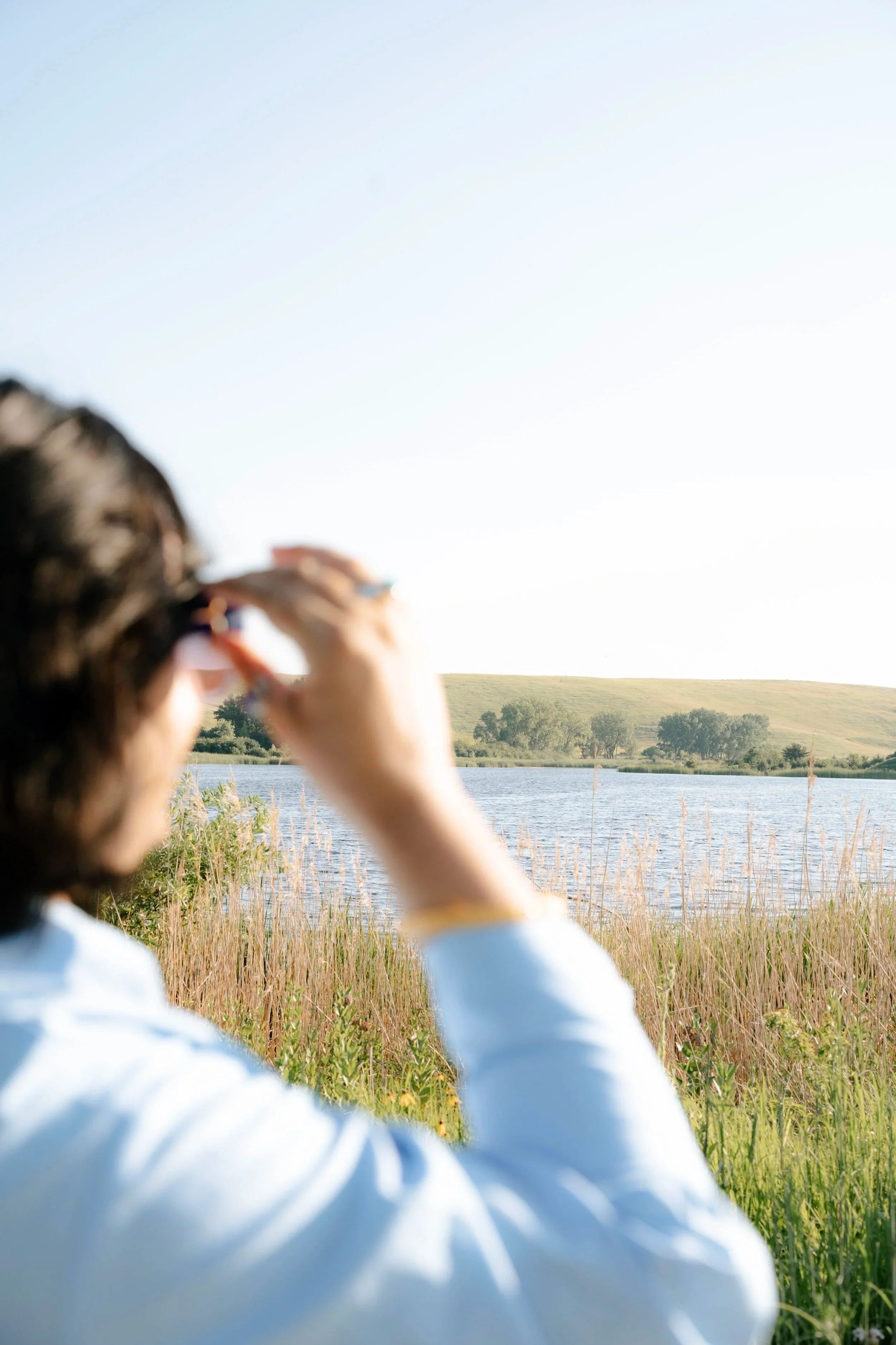 An image of the back of the head and arm of the author. She is out of focus, with her hand grabbing her sunglasses, looking into the fourground. The foreground is a beautiful pond with some hills behind it and tall prairie grass in front.
