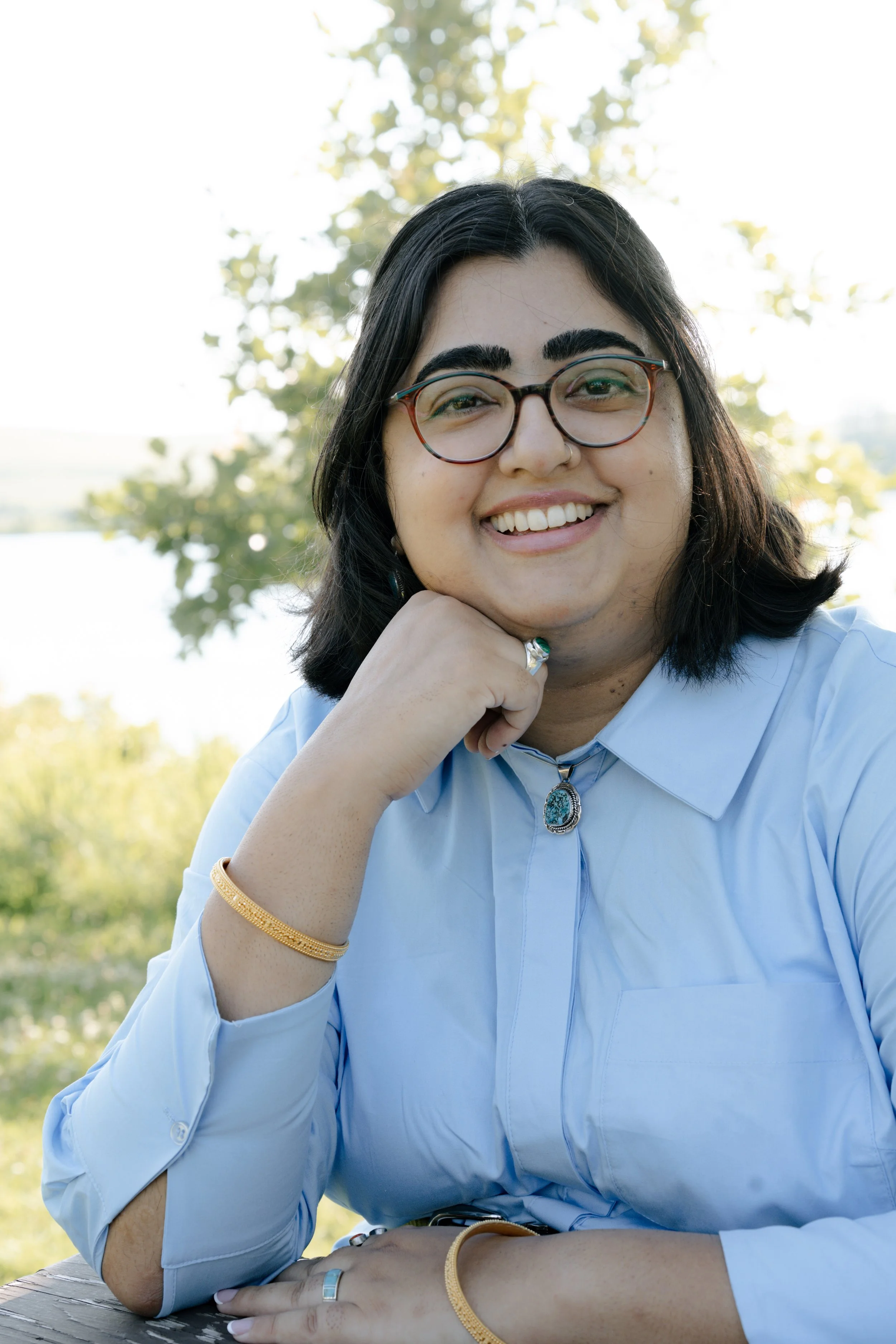 A brown woman with reddish-teal glasses is smiling at the camera with her fist under her chin. She is wearing gold bangles, a ring, a turquoise necklace and a blue collared shirt