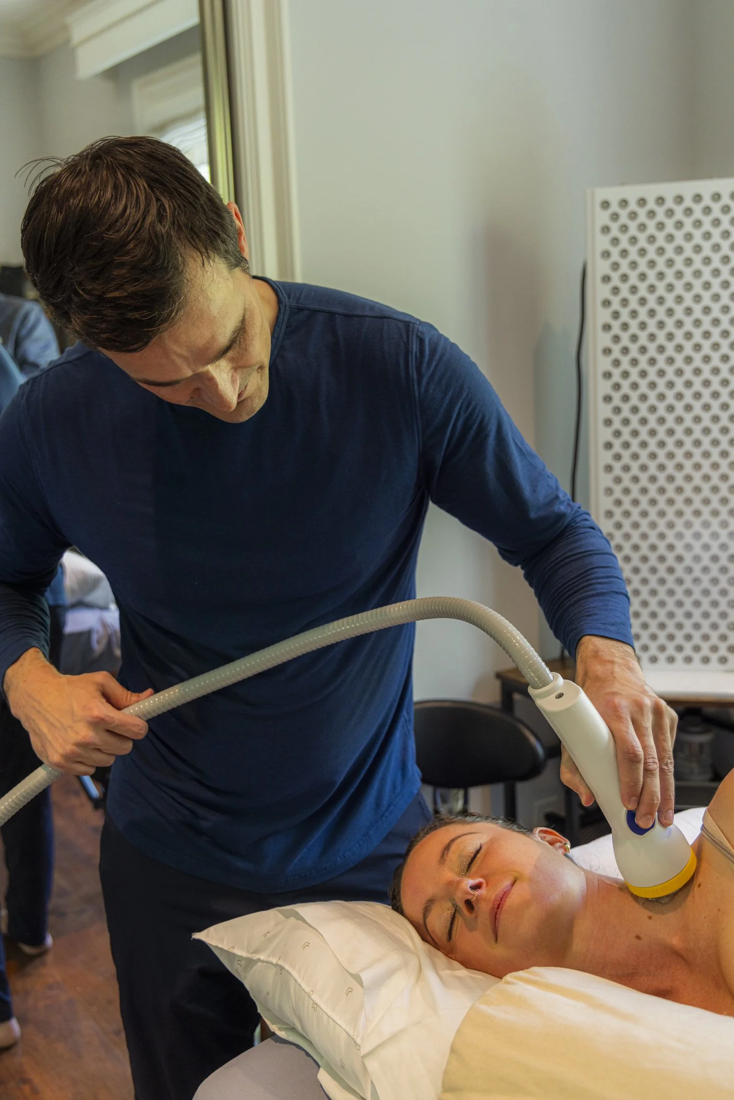 A woman relaxes with eyes closed while a healthcare provider uses light therapy device on her face in the medical setting of Biohacker Rehab.