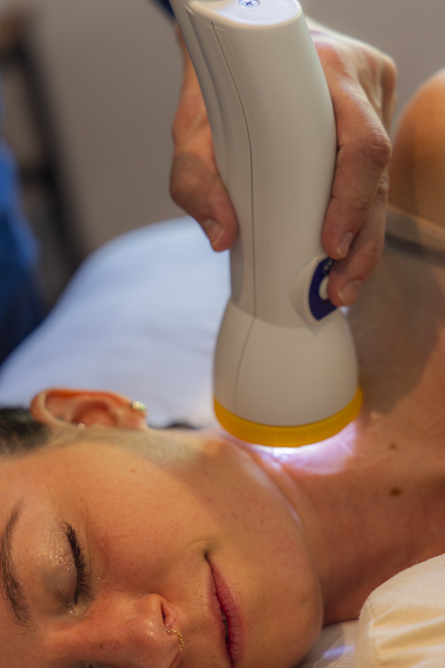A person with their eyes closed receiving a massage or treatment using a modern handheld device on their neck or shoulder area.
