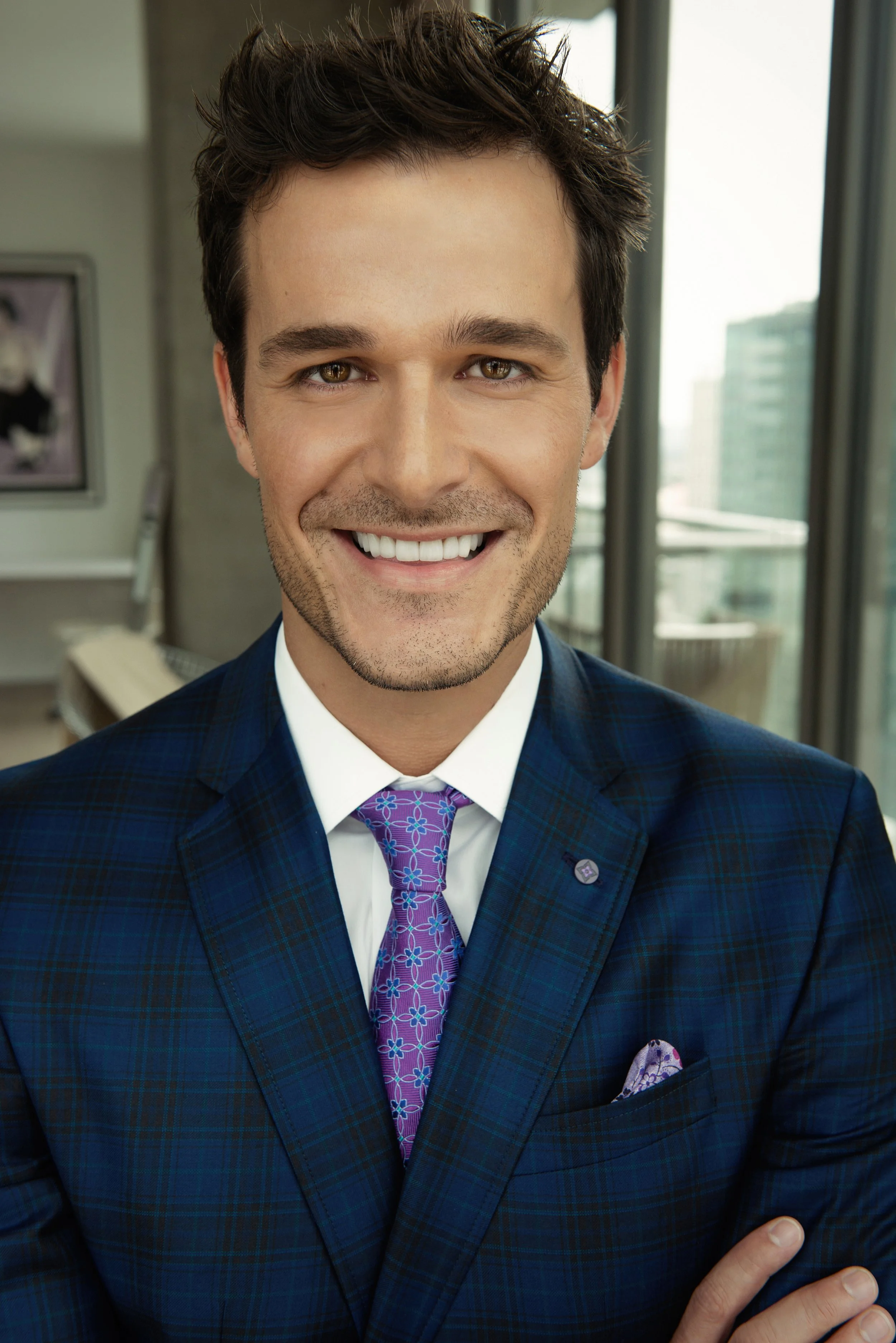 Close-up of a smiling man named Jimmy Clabots wearing a blue plaid suit, white shirt, purple floral tie, and matching pocket square, standing in an office with large windows and cityscape in the background.