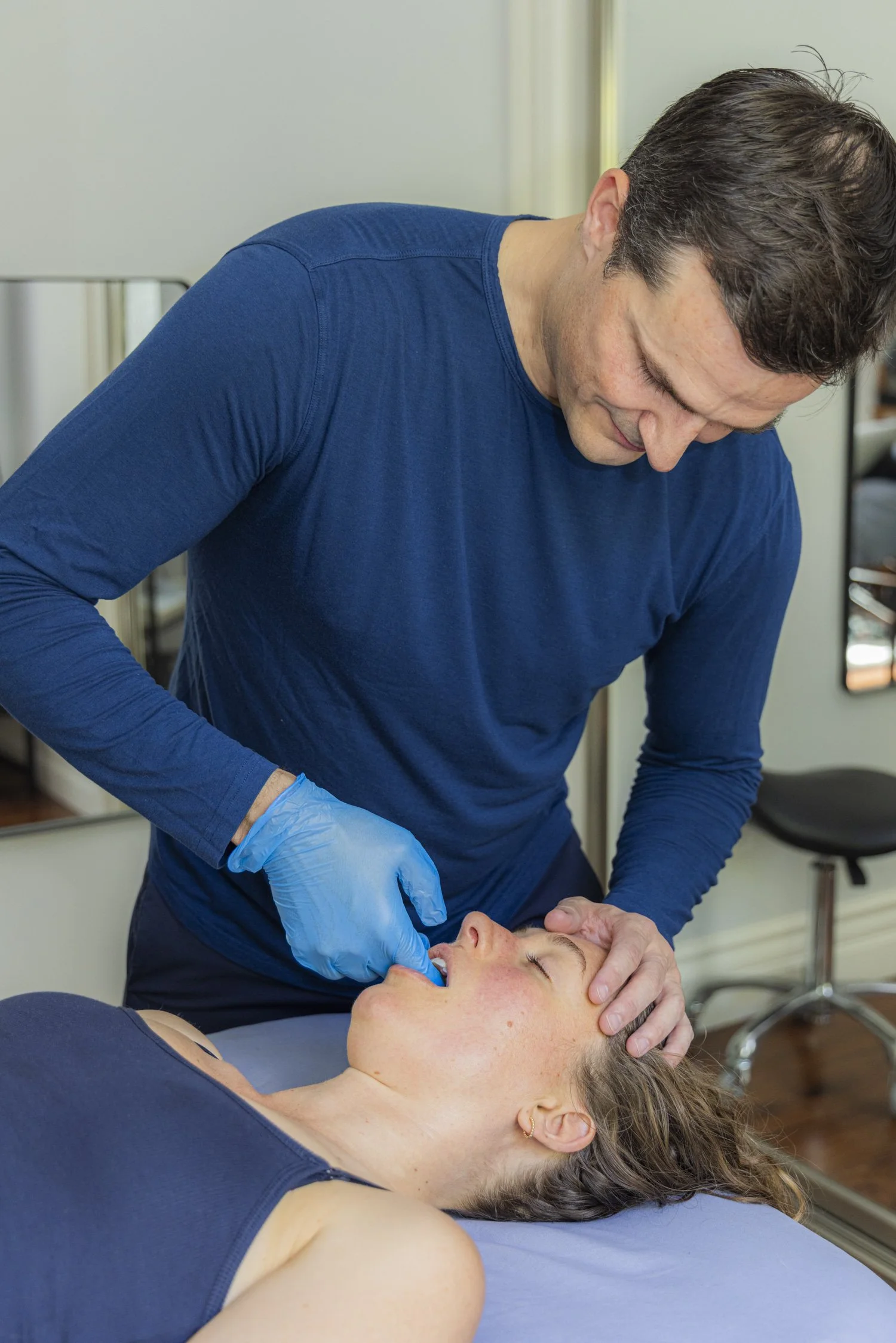 A man, Jimmy Clabots, is performing intra-oral CranioSacral Therapy on a woman lying down with her eyes closed, her head supported by a pillow, in a clinical setting.