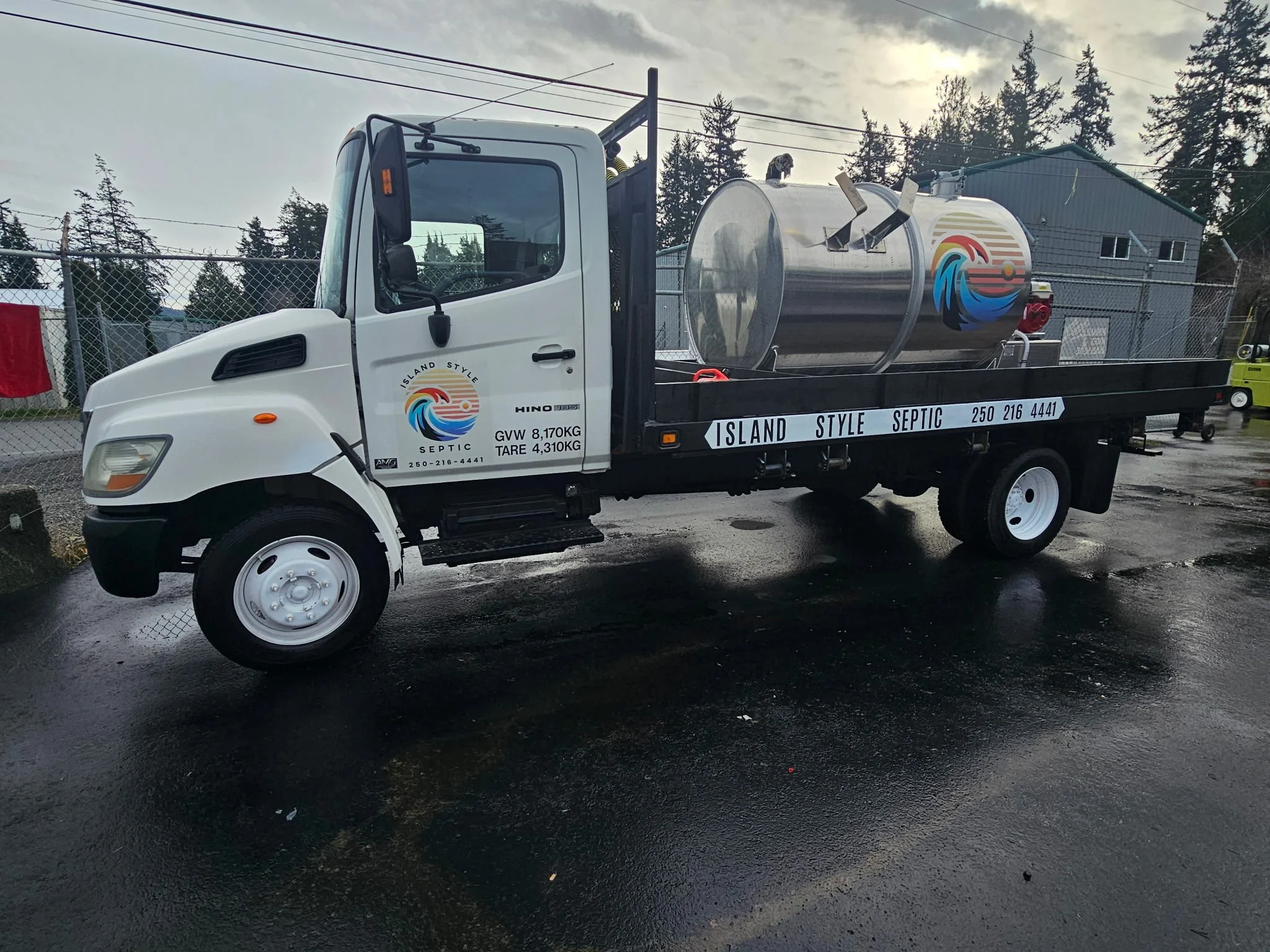 White flatbed truck branded with Island Style Septic, carrying a large cylindrical stainless steel septic tank with colorful wave logo, parked on wet asphalt.