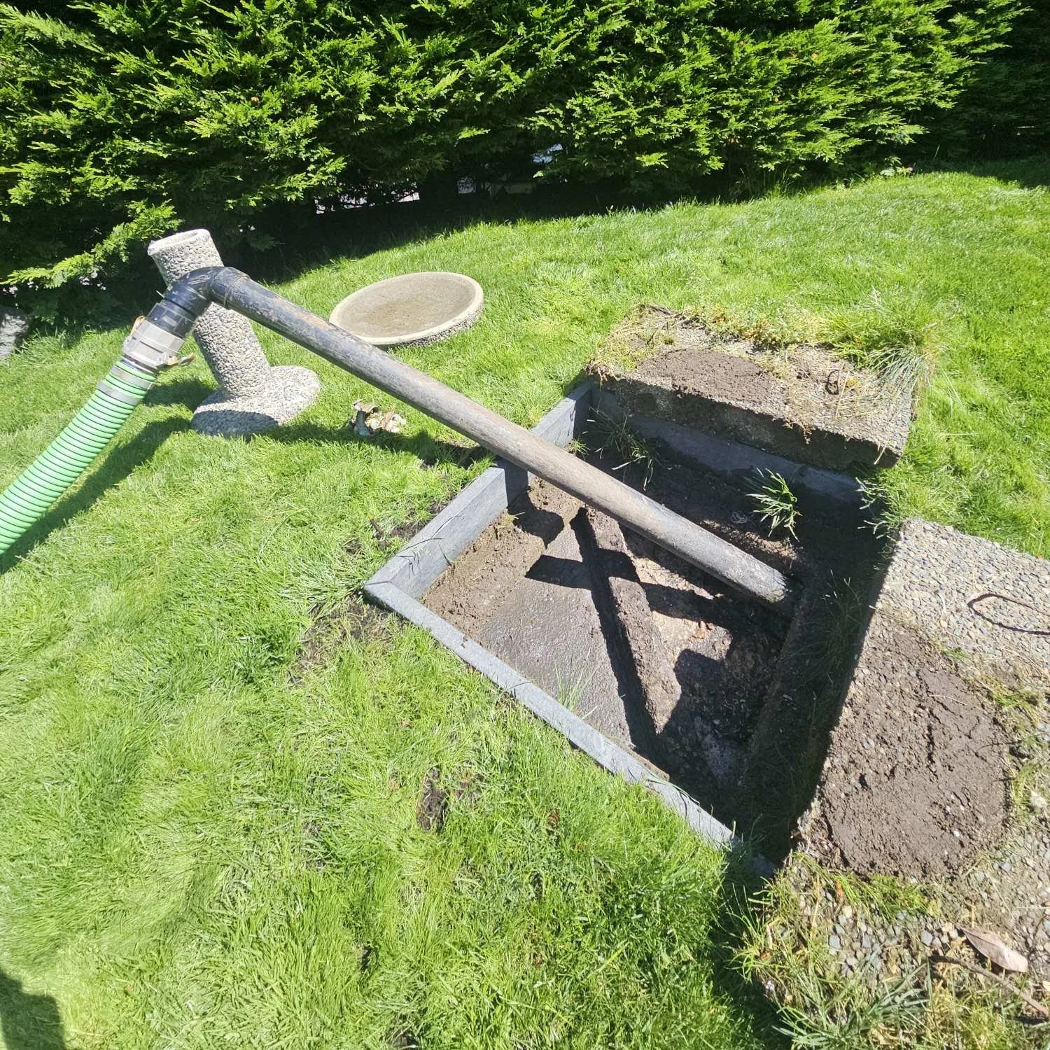 An open square hole in the ground with a wooden frame around it, and construction materials and tools nearby, in a grassy yard with bushes in the background.