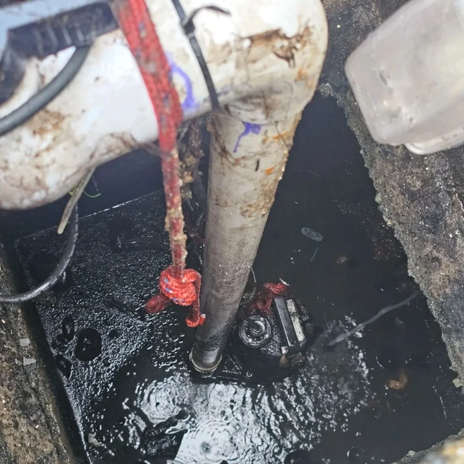 A close-up view of a municipal well or pump installed underground, with a pipe, electrical wires, and a red rope tied to the pipe, surrounded by wet soil and a layer of black liquid, possibly water or sludge, at the bottom.