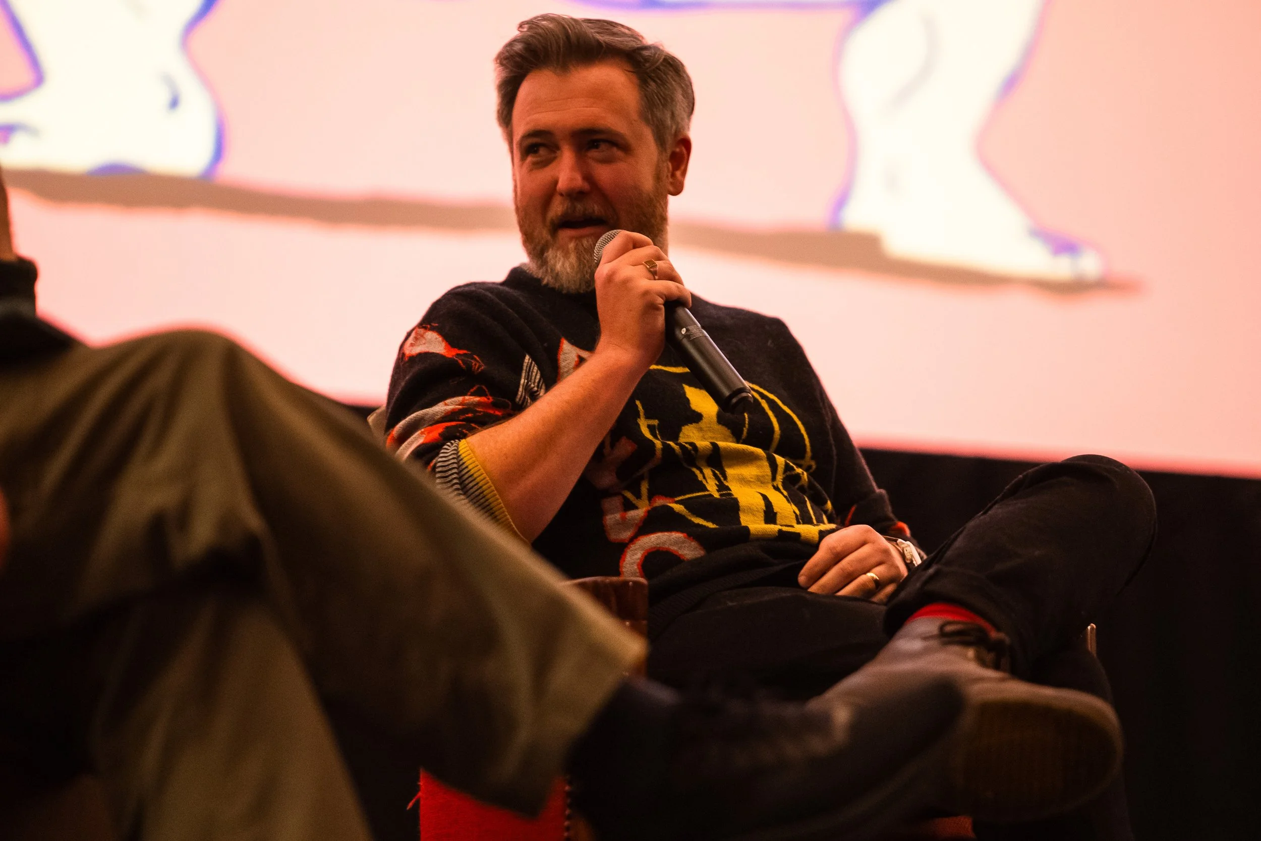 A man with a beard and styled hair is sitting on a chair, holding a microphone near his mouth. He is wearing a black sweater with a colorful graphic design. In the background, there is a large pink screen with abstract line art. The setting appears t