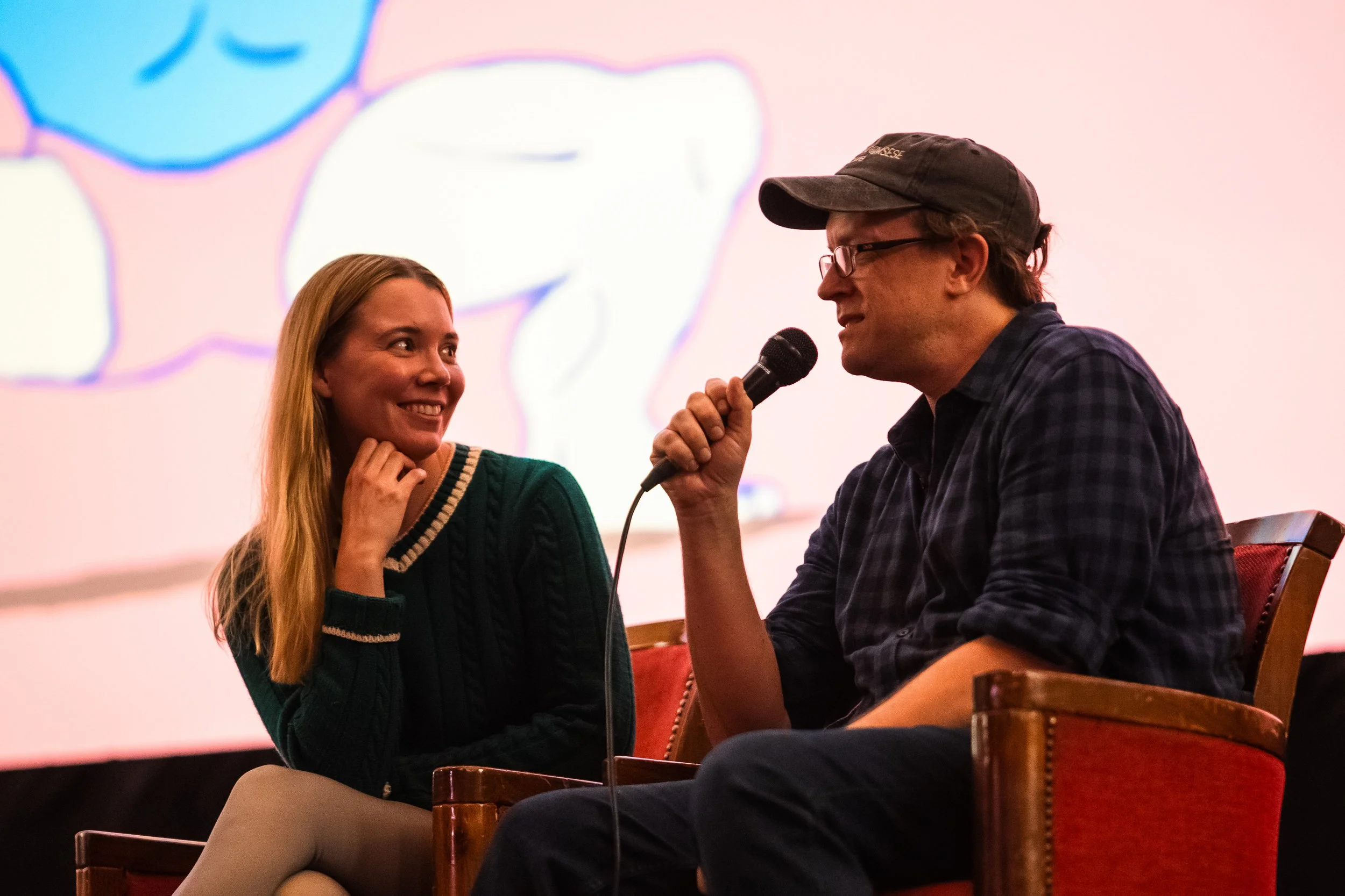A young woman and a man are sitting and engaging in a conversation, with the man holding a microphone. They are indoors, sitting on wooden chairs, with a bright background displaying a large, stylized cartoonish face.