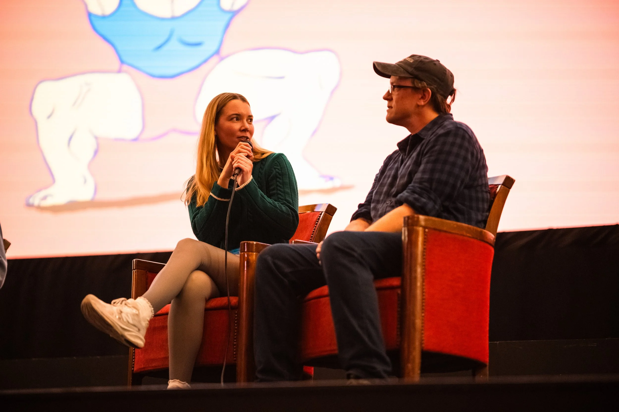 A woman with long blonde hair in a green sweater, beige stockings, and white sneakers is holding a microphone and speaking into it. She is sitting on a red upholstered chair on stage, looking at a man with glasses, a black cap, and a dark checkered s