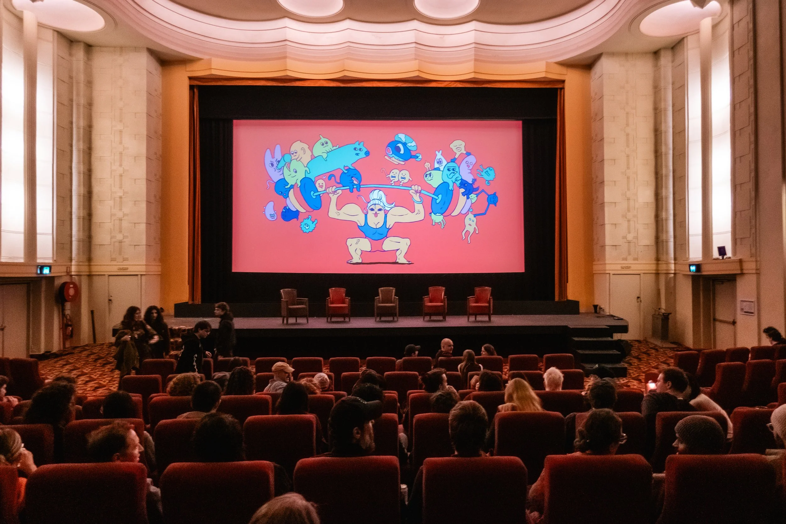An auditorium with a large screen displaying a colorful drawing of a muscular figure lifting a barbell with various cartoon-like animals and creatures on or around it. There are empty chairs on stage and several people seated and standing in the audi