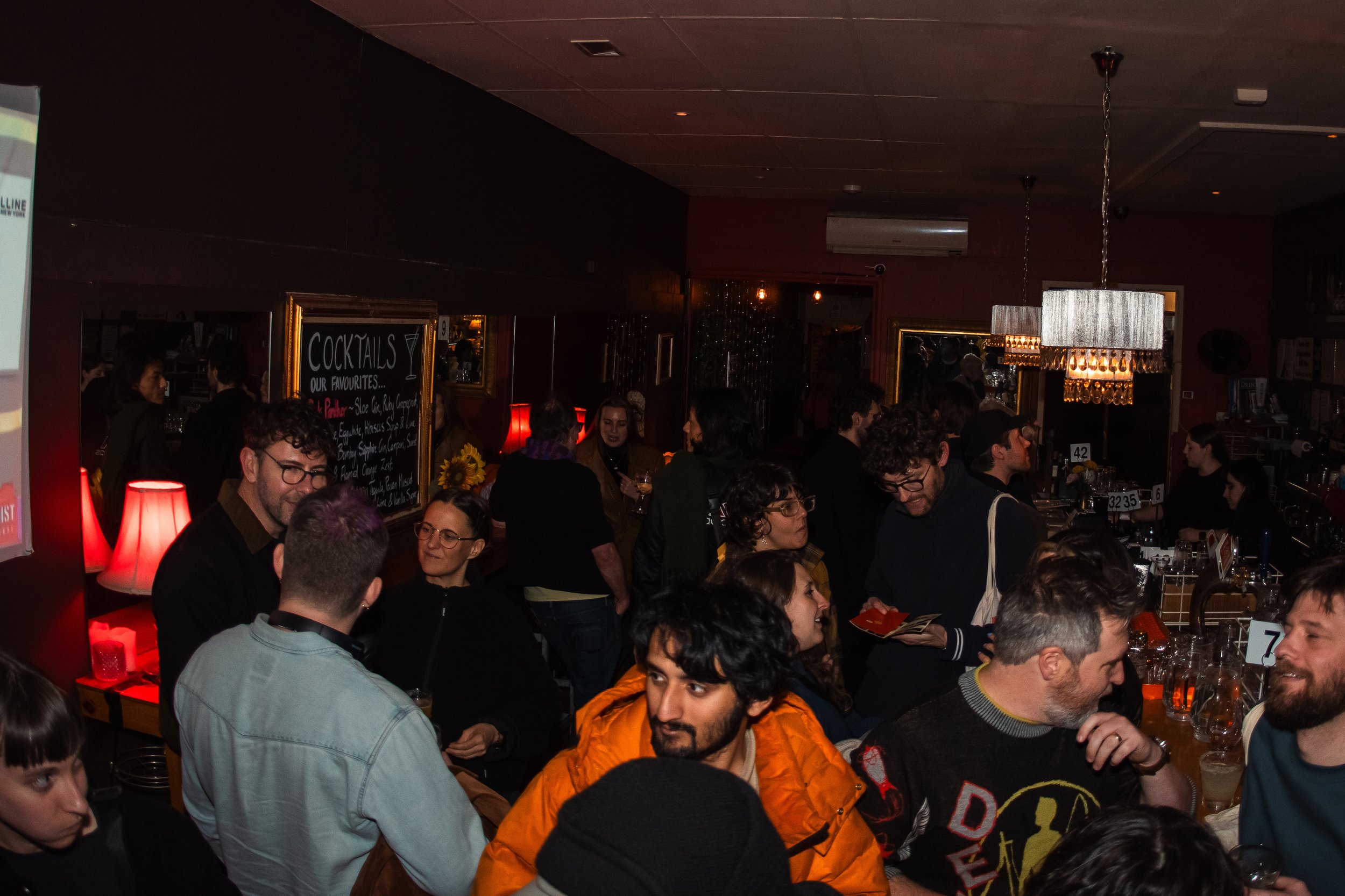 A crowded bar or restaurant with people socializing, some holding drinks. There are hanging light fixtures, a chalkboard menu for cocktails, and a red lamp on a table.