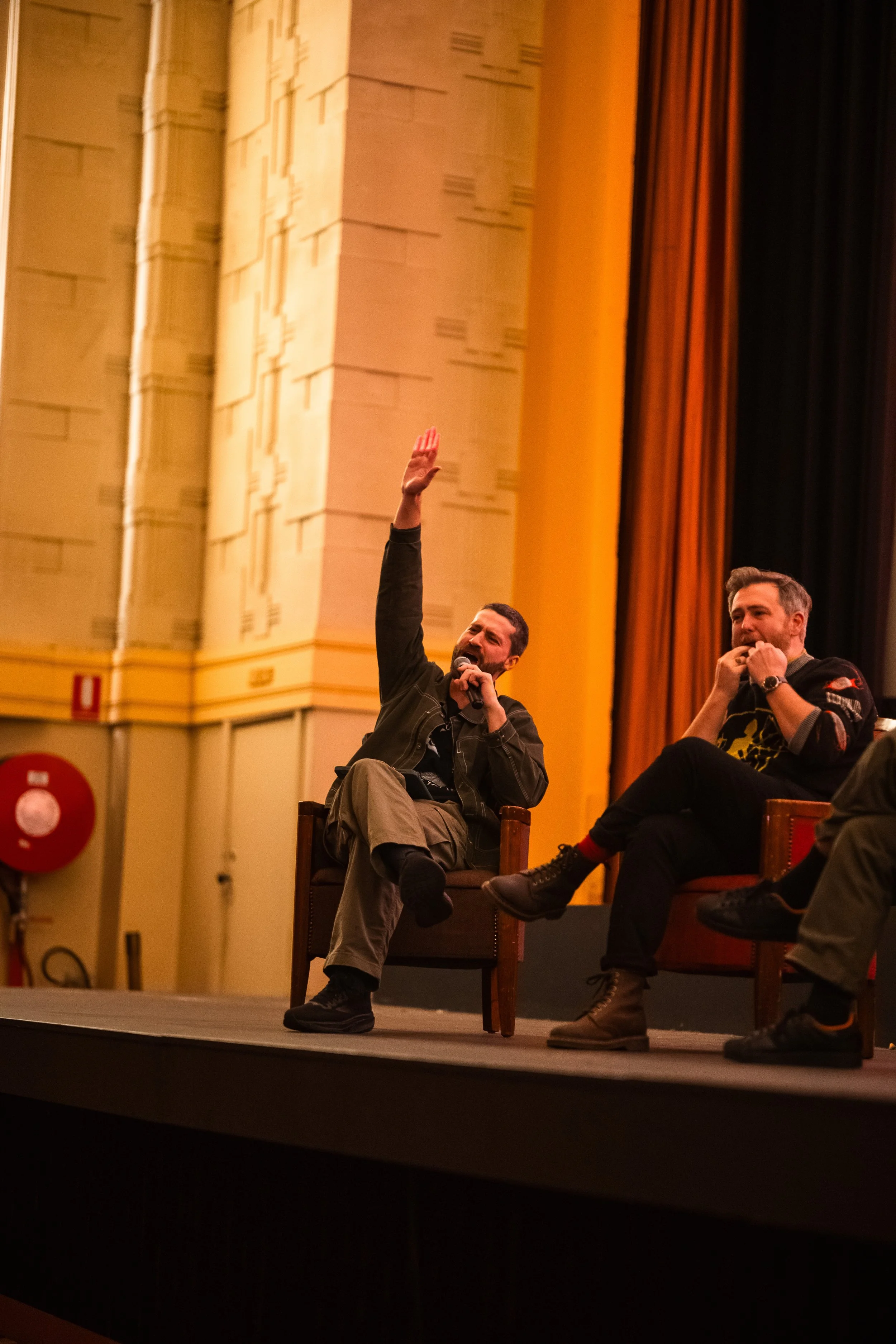 Two men are sitting on chairs on a stage, one holding a microphone with his hand raised and the other with his hand near his mouth, in an auditorium with curtains and a decorative wall.