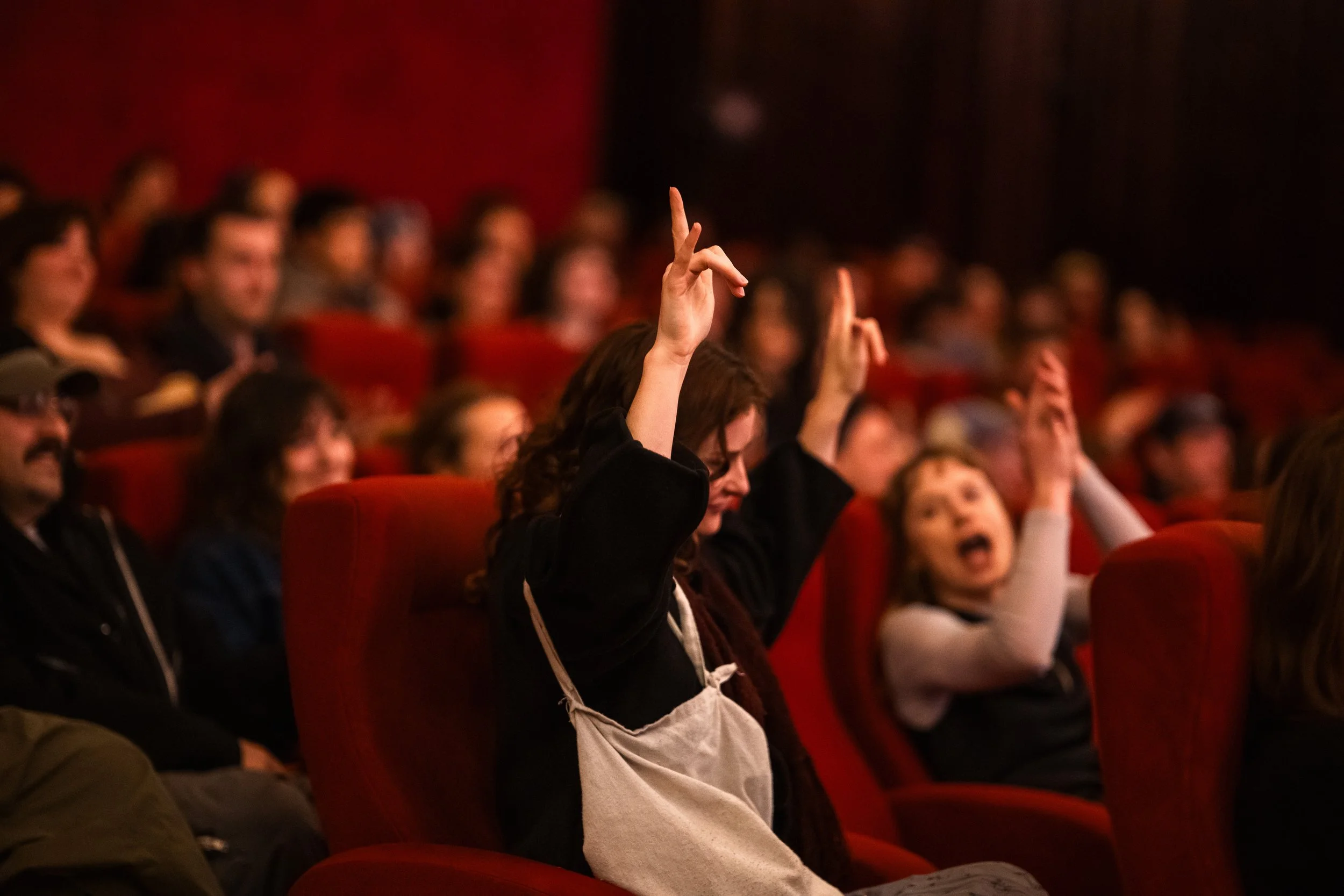 Audience at a conference or cinema, with a woman in the front raising her hand to ask a question or make a comment.