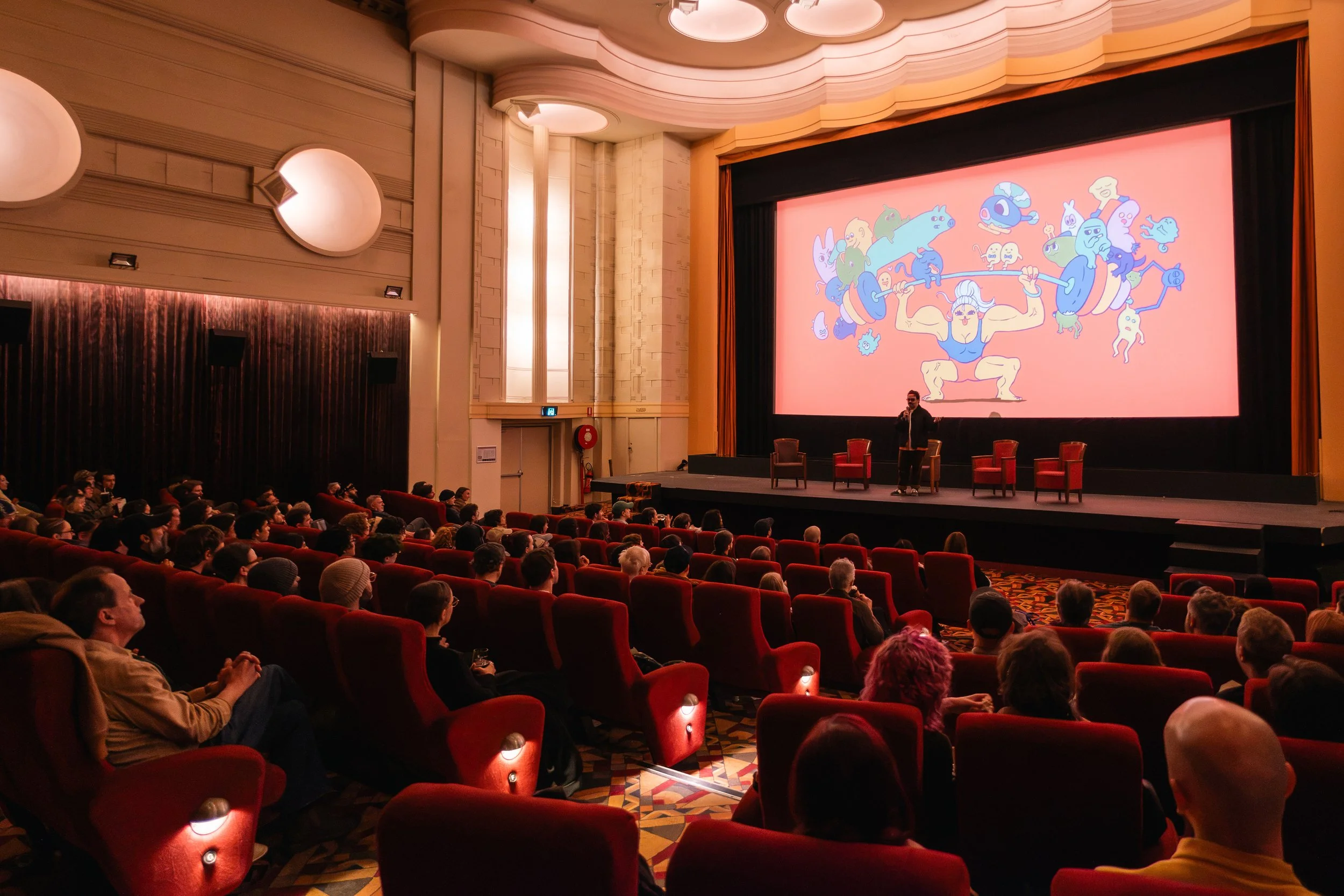 Audience watching a presentation in a theater with a large screen displaying a cartoon muscular character lifting weights, surrounded by various colorful cartoon creatures.