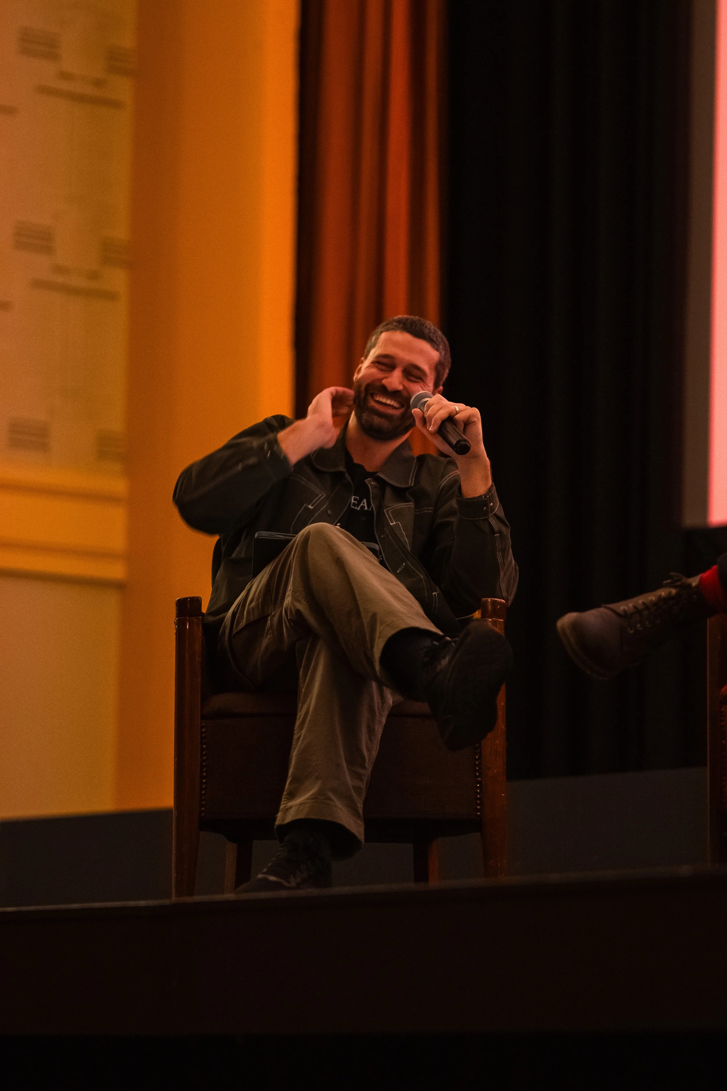 A man with a beard smiling while holding a microphone and sitting on a chair on a stage with black curtains and warm-colored walls.
