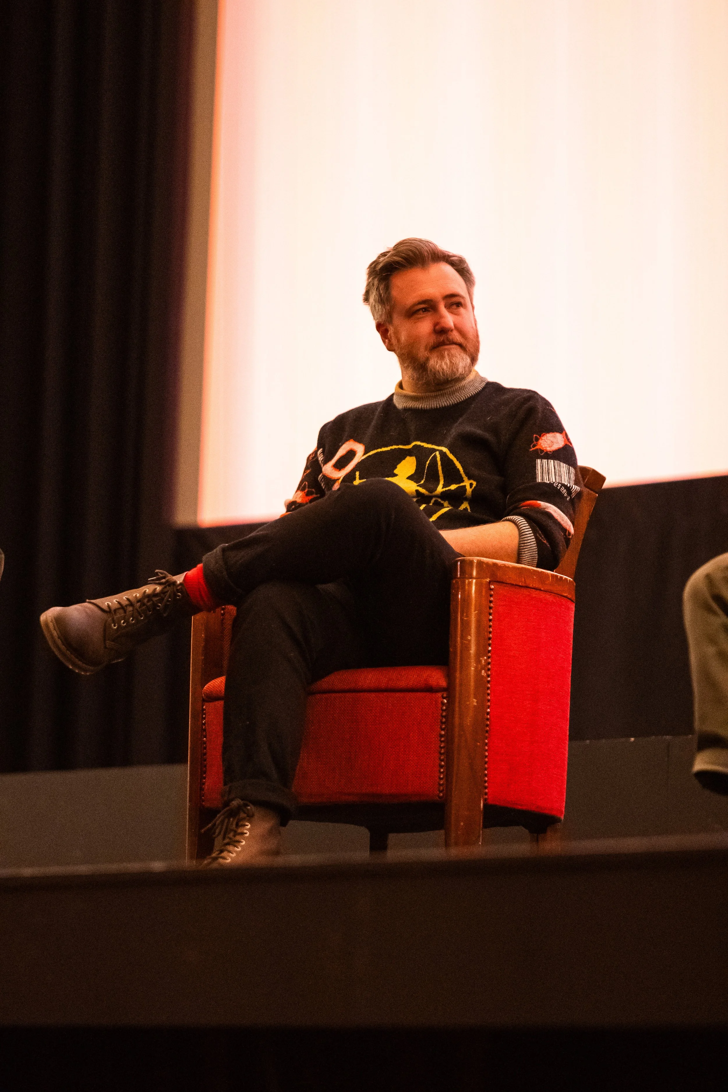 A man with a beard and gray hair sitting in an orange chair on a stage during an event or panel, wearing a black sweater with colorful designs, and black pants, crossed legs, and moccasin-style shoes, with a neutral expression.