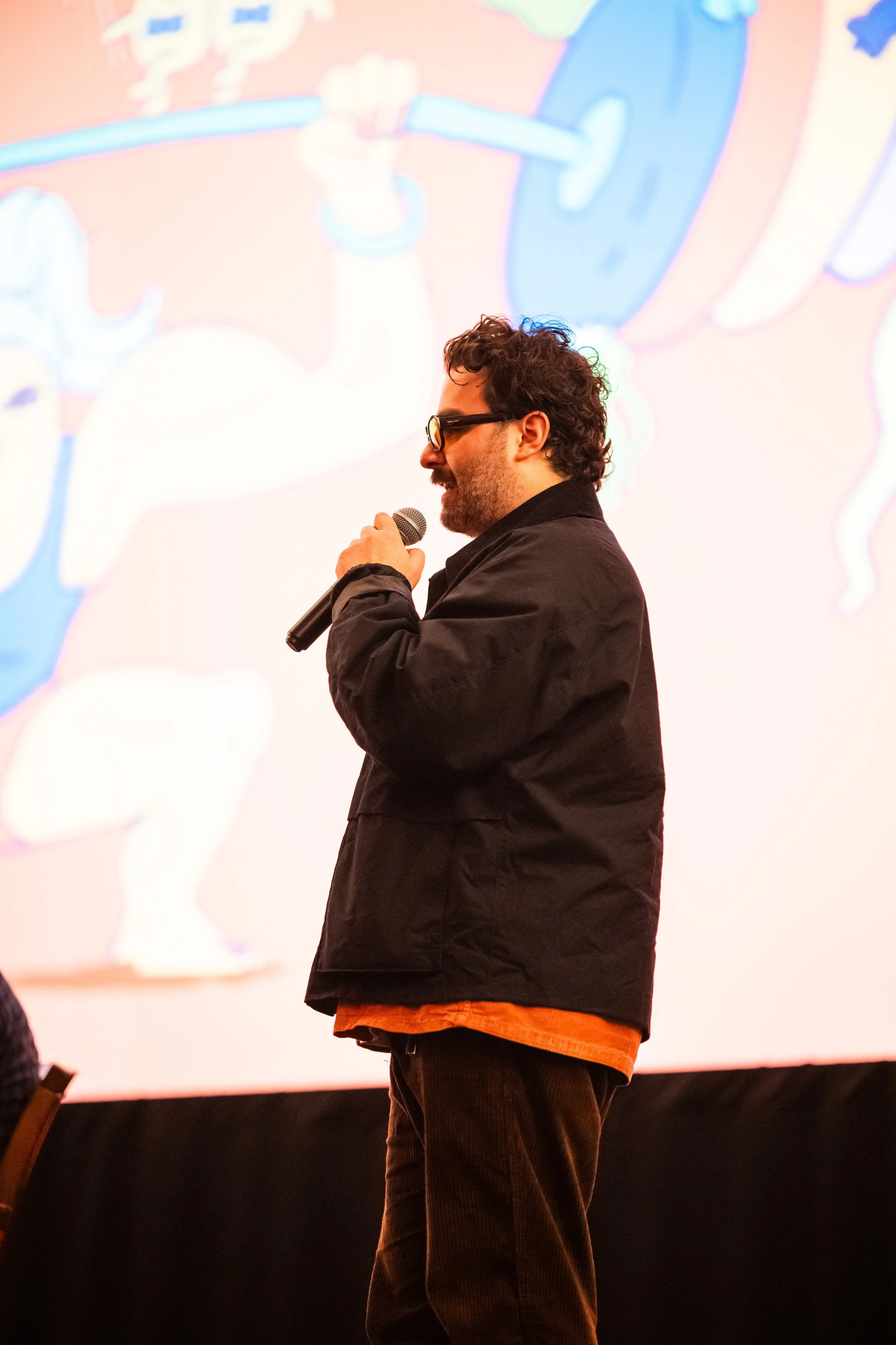 A man with dark curly hair and glasses speaking into a microphone at an event, with a colorful abstract background projected behind him.