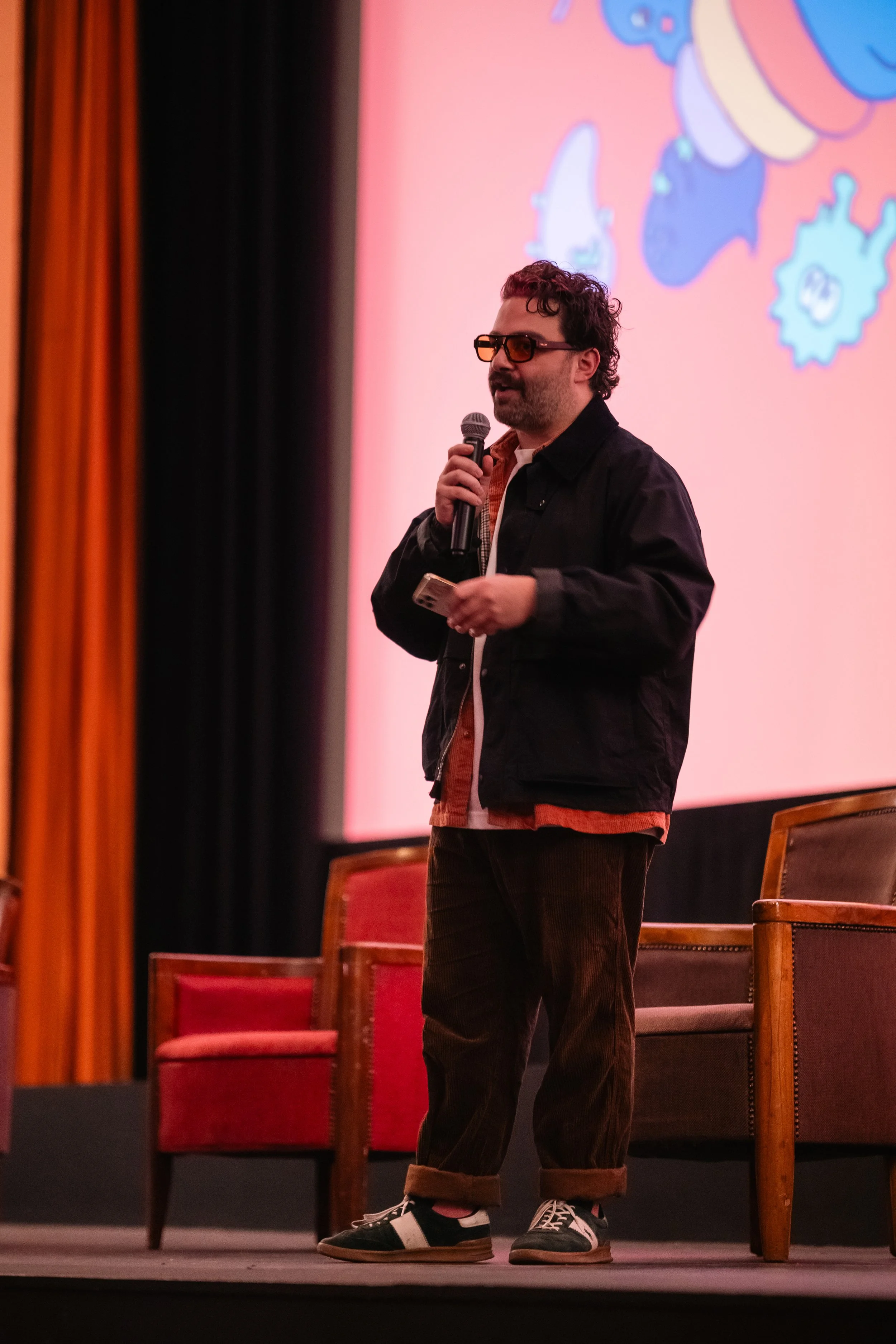A man with curly hair, beard, and glasses holding a microphone and a phone standing on stage in front of a pink background with cartoon gear illustrations. He wears a black jacket, plaid shirt, dark pants, and sneakers with white laces.