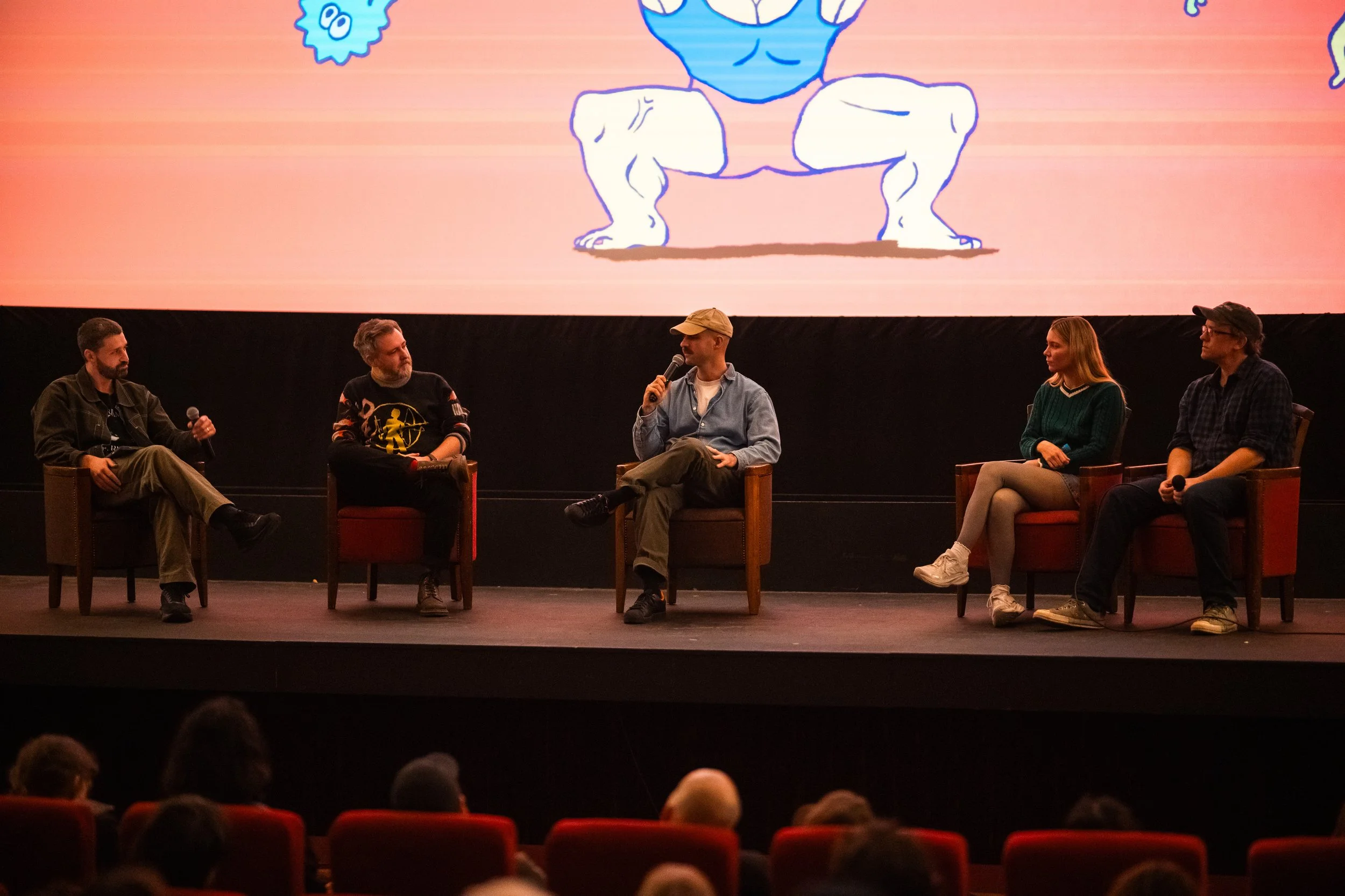 Panel discussion with five people seated on stage, one holding a microphone, in front of a large screen displaying a cartoon character lifting weights.