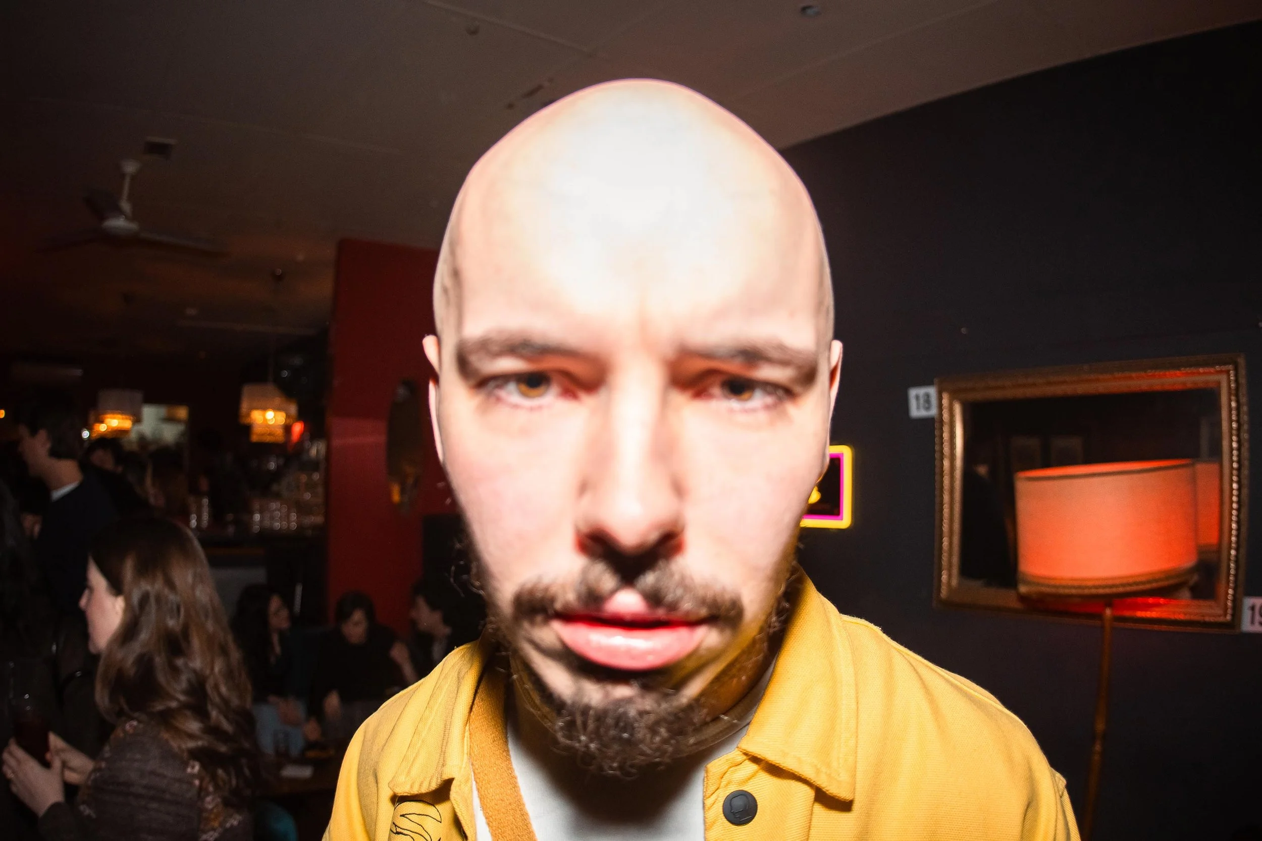 Close-up of a man with a bald head, brown eyes, and a beard, wearing a mustard yellow jacket, in a dimly lit indoor setting with people and framed pictures in the background.