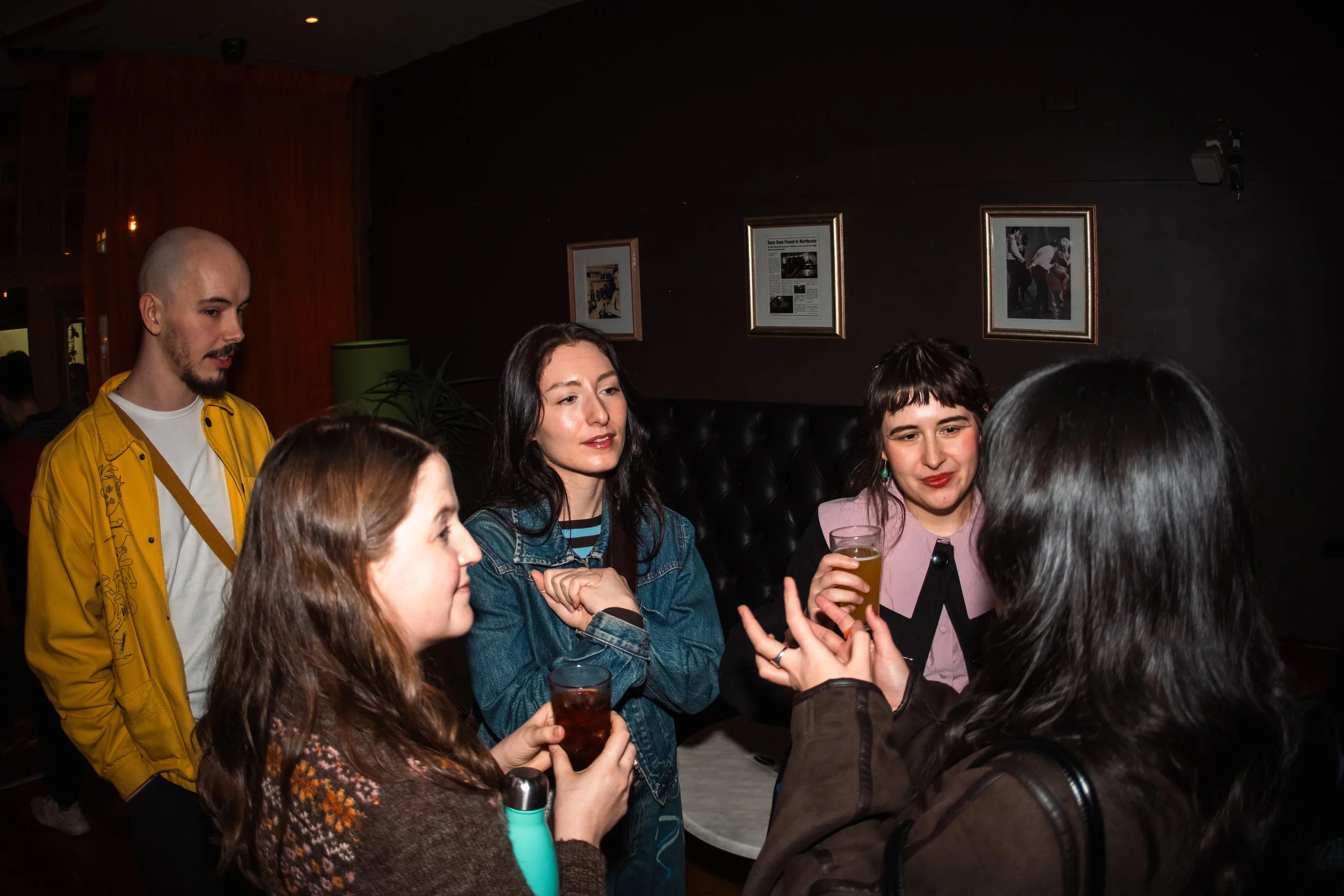 Group of people socializing at a bar or nightclub, with some holding drinks, in a dimly lit setting with framed pictures on the wall.