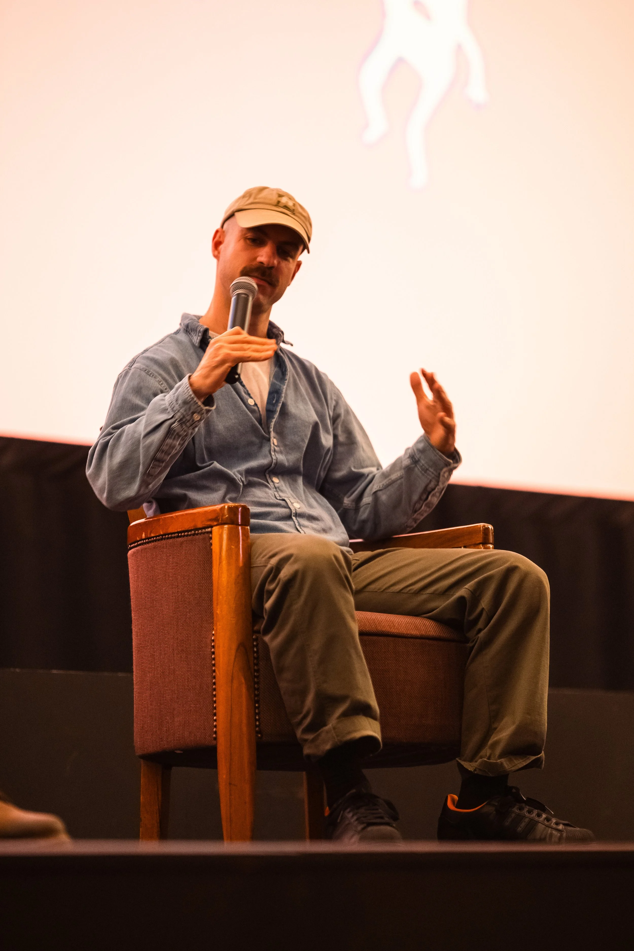 A man sitting in a chair on stage holding a microphone, wearing a baseball cap, denim shirt, khaki pants, and black sneakers with orange accents. There is a pink and white neon horse outline projected on the wall behind him.