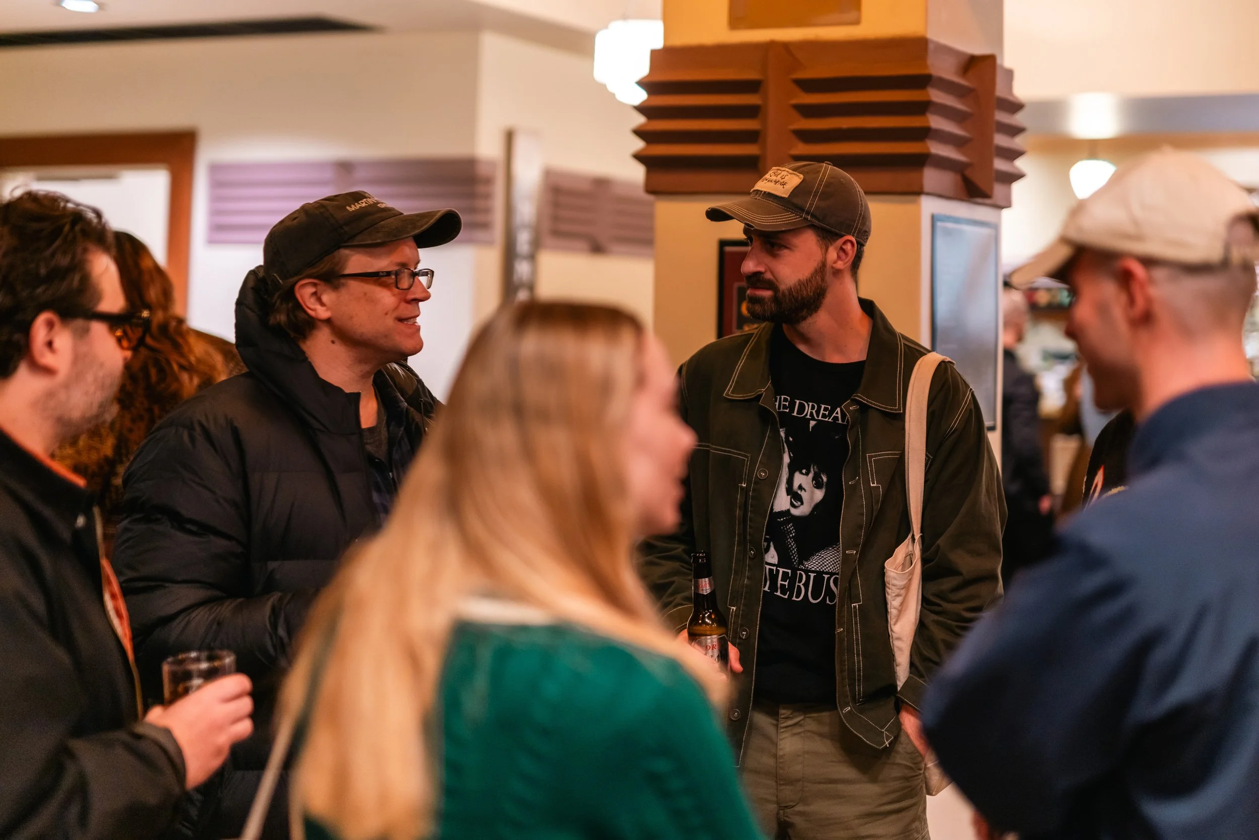 People socializing at an indoor gathering, some holding drinks, with a focus on two men engaged in conversation, one wearing a black cap and glasses, the other wearing a gray cap and a black graphic t-shirt.