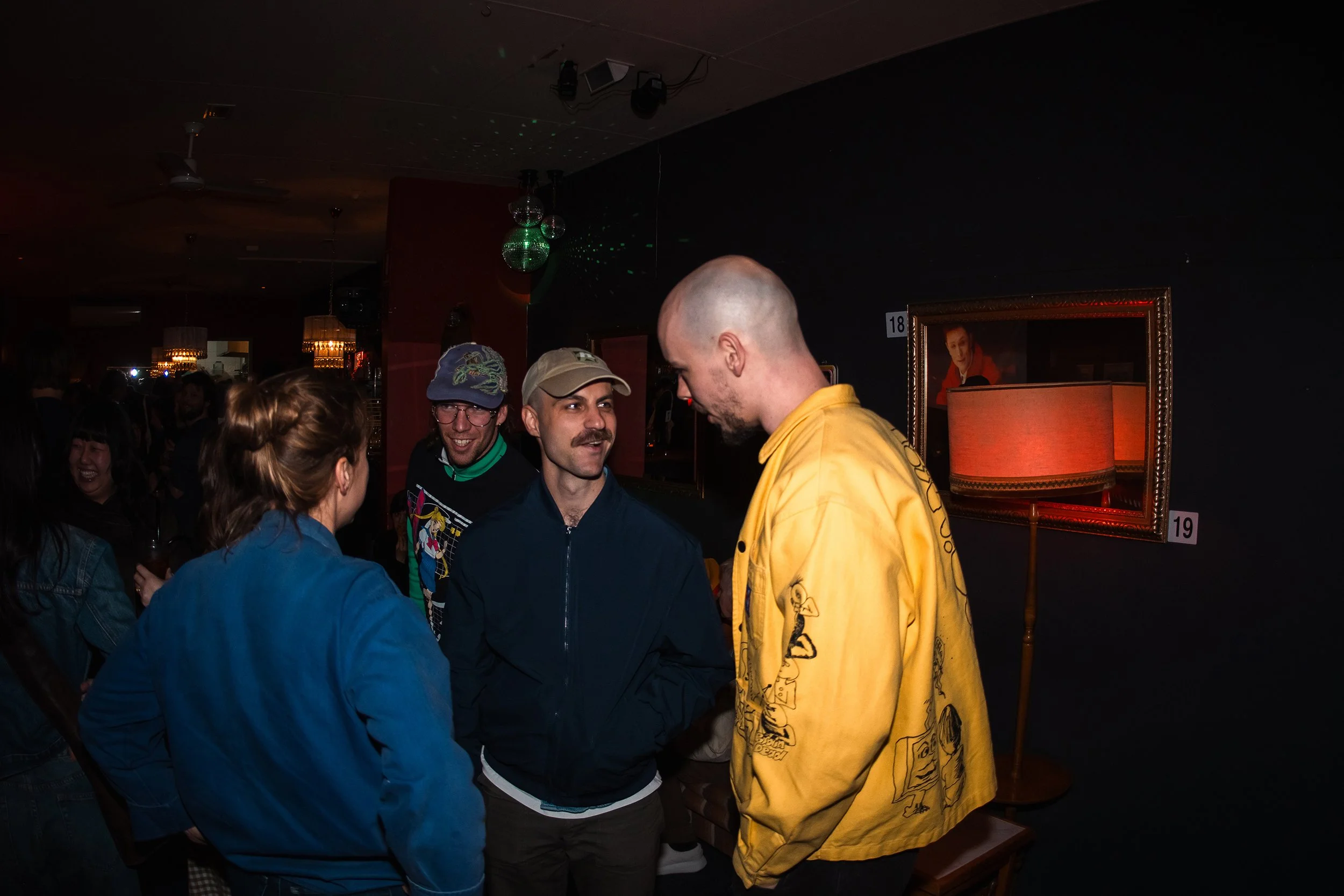 A group of people socializing at an indoor event or party, with some people facing each other and smiling. The room features dark walls, framed pictures, and a lamp, and there are colorful disco lights projected from the ceiling.