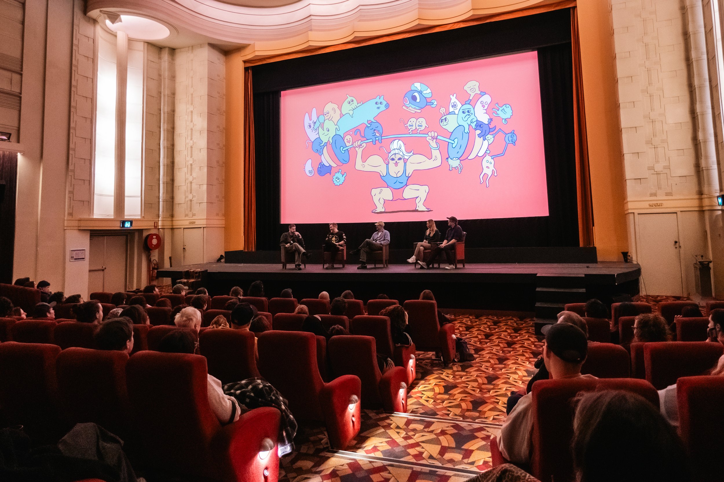 A theater auditorium with a stage where four people are seated in chairs, engaged in a panel discussion. The large screen behind them displays a colorful cartoon illustration of a muscular woman lifting a barbell with various cartoon characters aroun