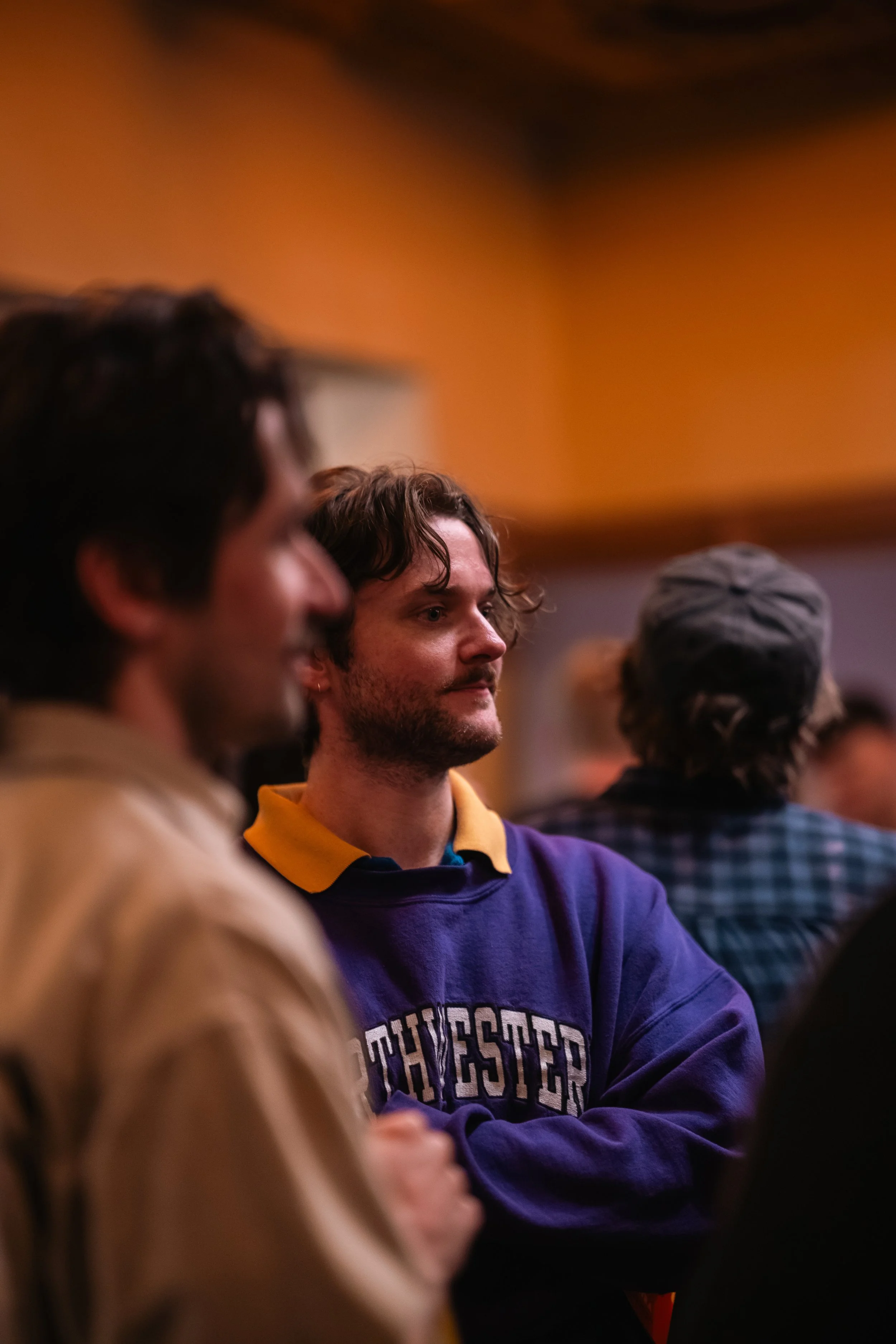Group of young people sitting together in a room with warm lighting, one person in focus wearing a purple sweatshirt with yellow collar and Oprah-Bridgeport written on it, others blurred in the background.