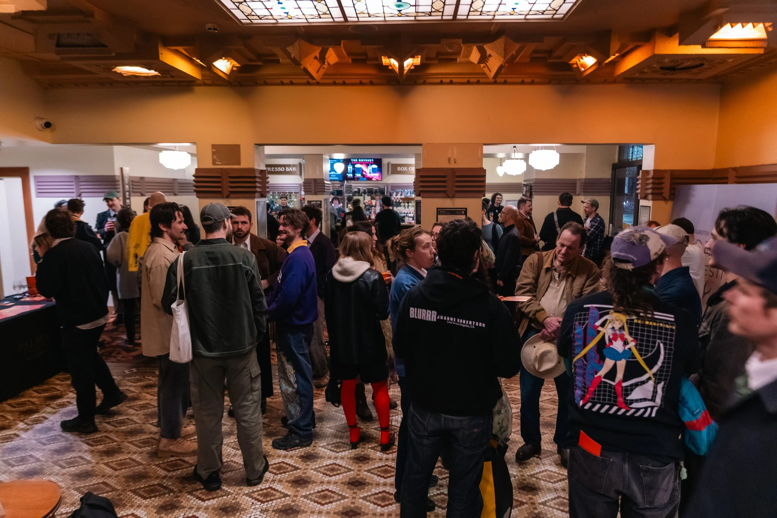 Crowd of people gathered inside a hall, engaged in conversation. There are food and drink counters in the background, illuminated signs, and decorative lighting fixtures hanging from the ceiling.
