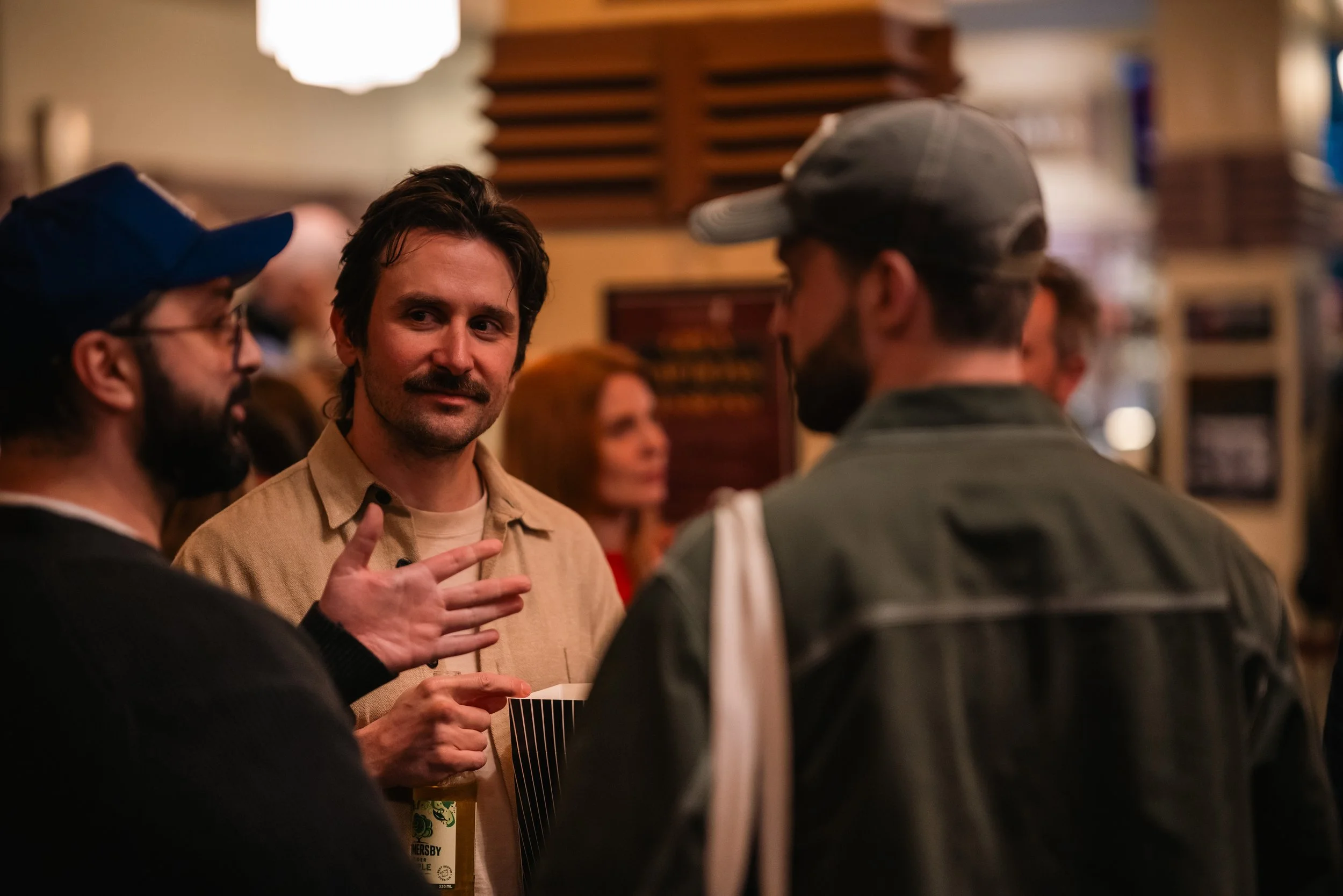 Group of people socializing indoors, including a man with dark hair and a mustache talking to another man wearing a cap, with others visible in the background.