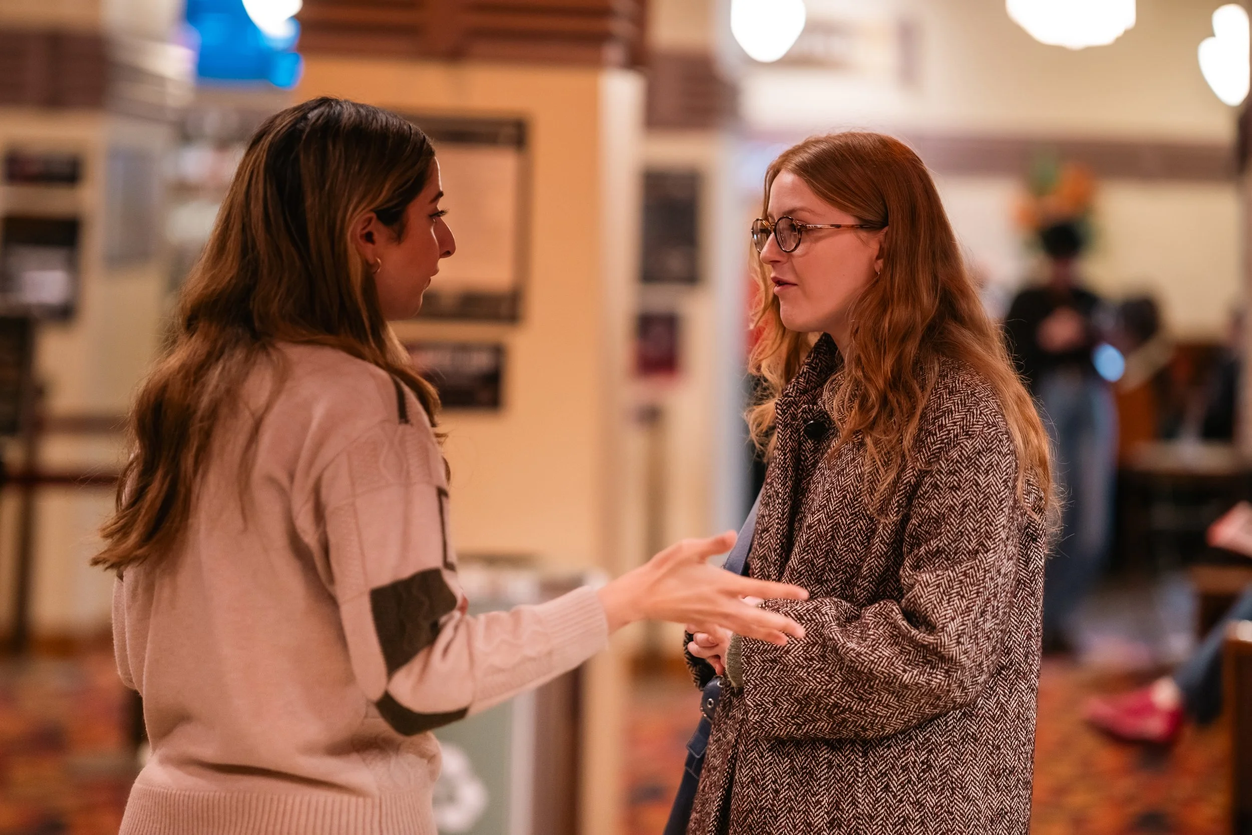 Two women are engaged in conversation indoors, with one woman in a light pink sweater and the other in glasses and a brown patterned coat, in a warmly lit room with a blurred background.