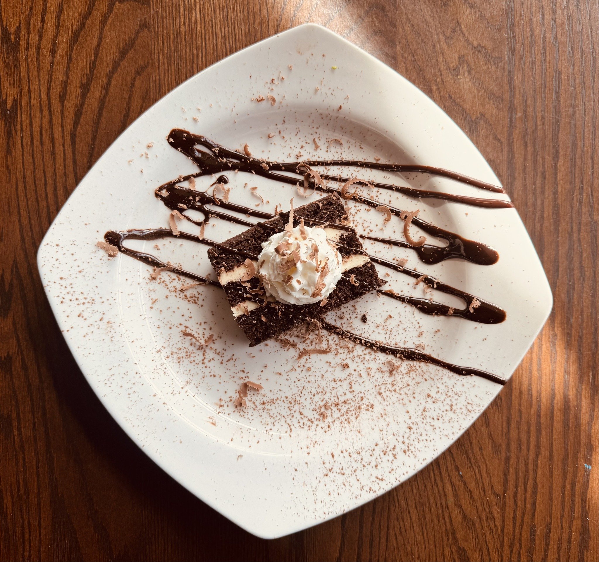 Chocolate brownie dessert with whipped cream, chocolate shavings, and chocolate drizzle on a white square plate placed on a wooden table.