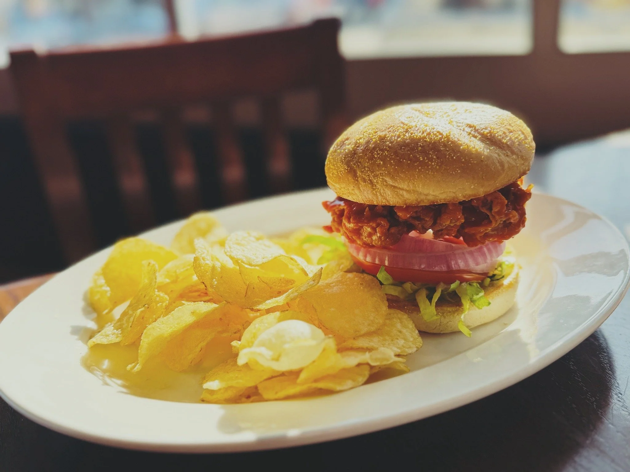 A plate with a fried chicken sandwich topped with pickles, onions, and lettuce, alongside potato chips, on a wooden table near a window.