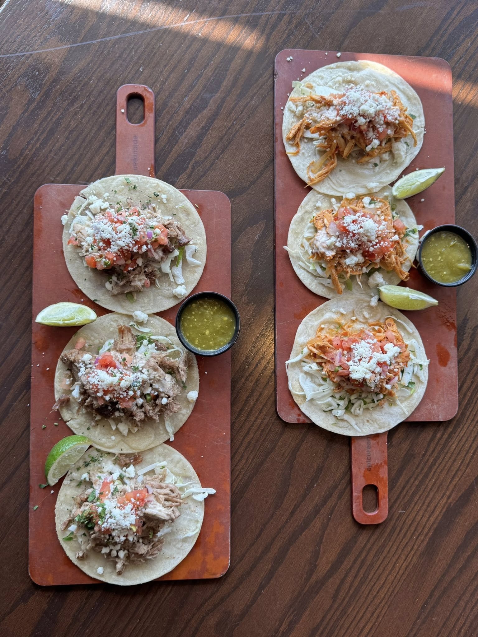 Two wooden trays each holding three tacos with shredded meat, salsa, cheese, and lime wedges, along with small cups of green sauce, on a wooden table.