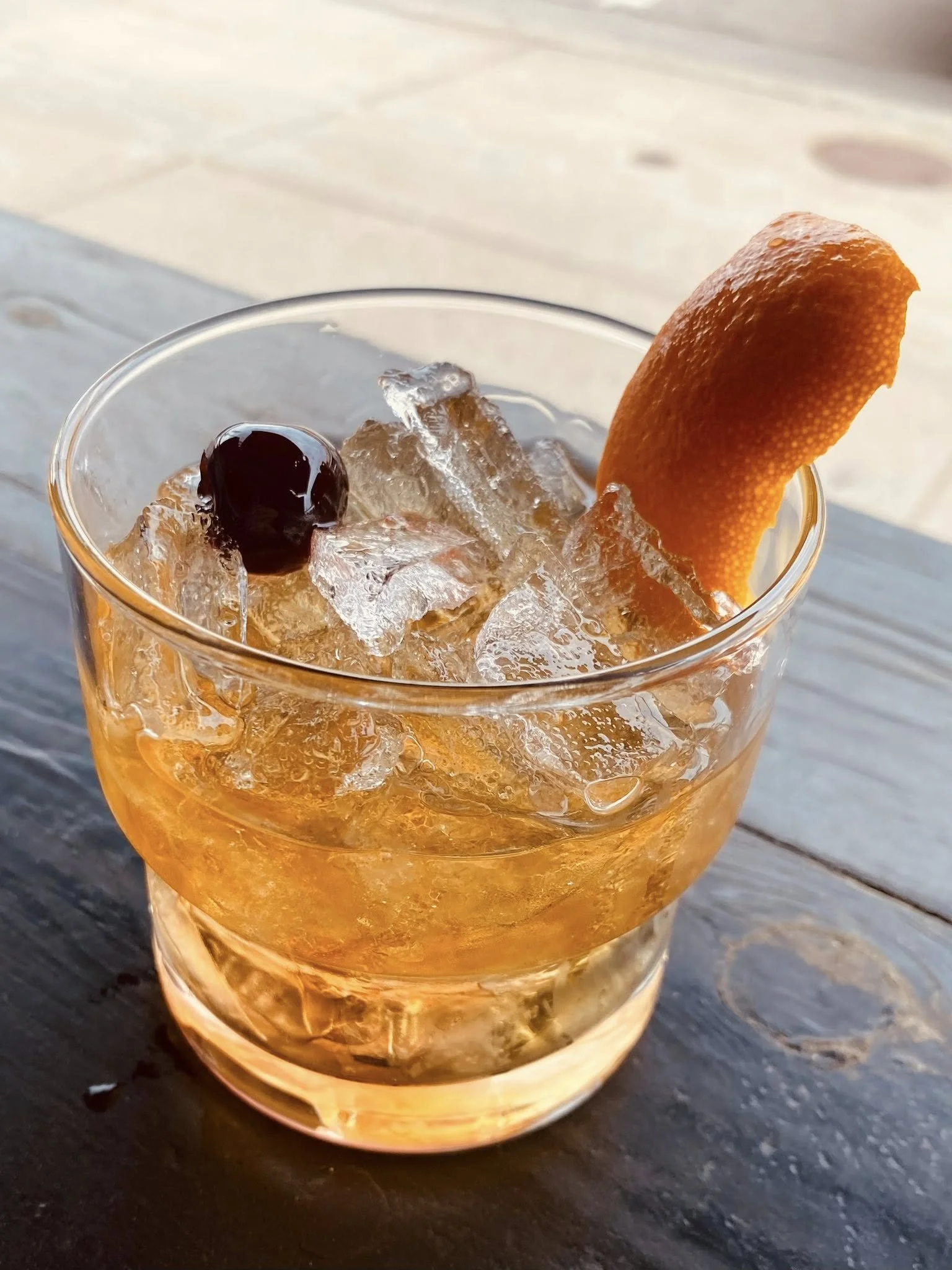 A glass of cocktail with ice, a cherry, and an orange twist garnish on a wooden surface.