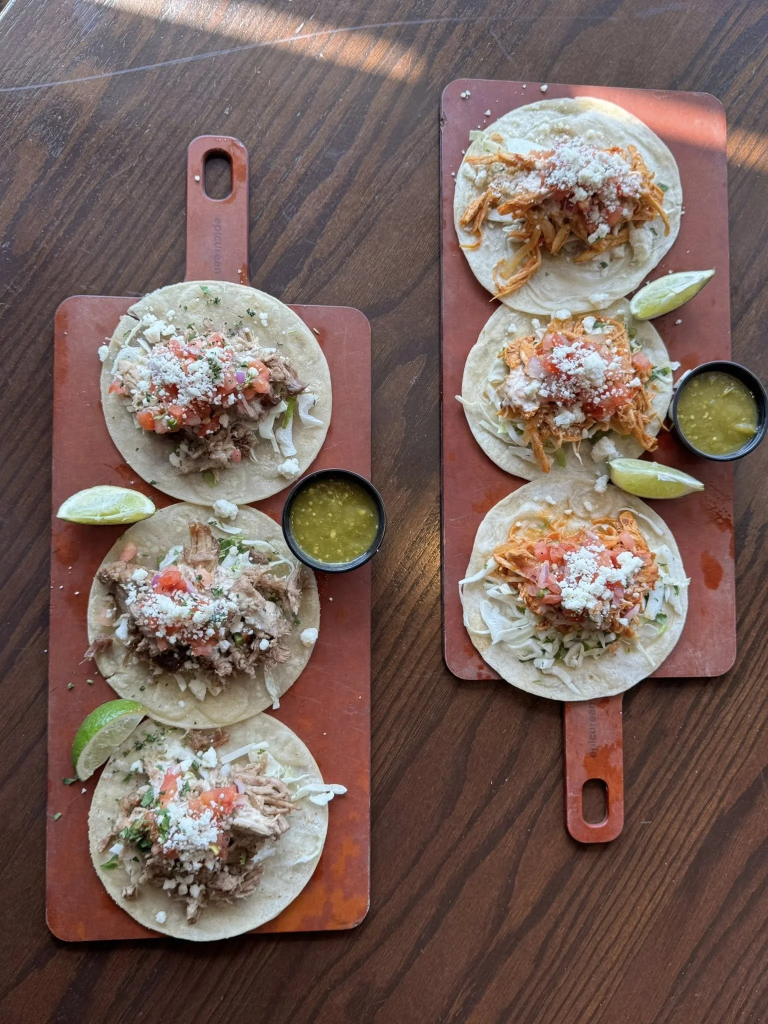 Two wooden serving boards each with three tacos, topped with shredded meat, shredded chicken, and shredded pork, with lime wedges and small bowls of green salsa.