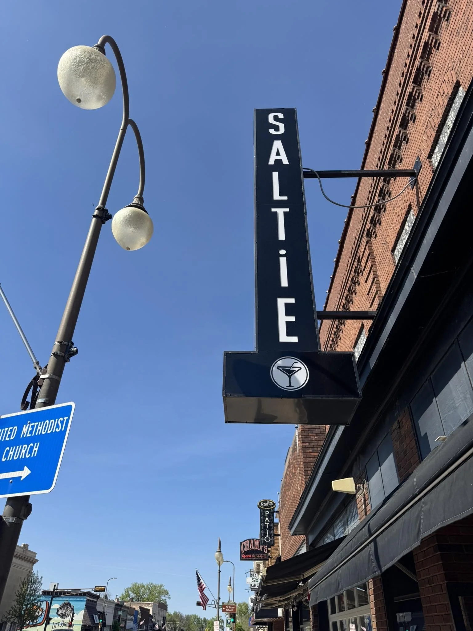 Street view showing a tall vertical sign for a bar called 'Saltie' with a martini glass icon, alongside a streetlamp with two round lights, a blue directional sign for United Methodist Church, and other storefront signs in the background on a clear day.