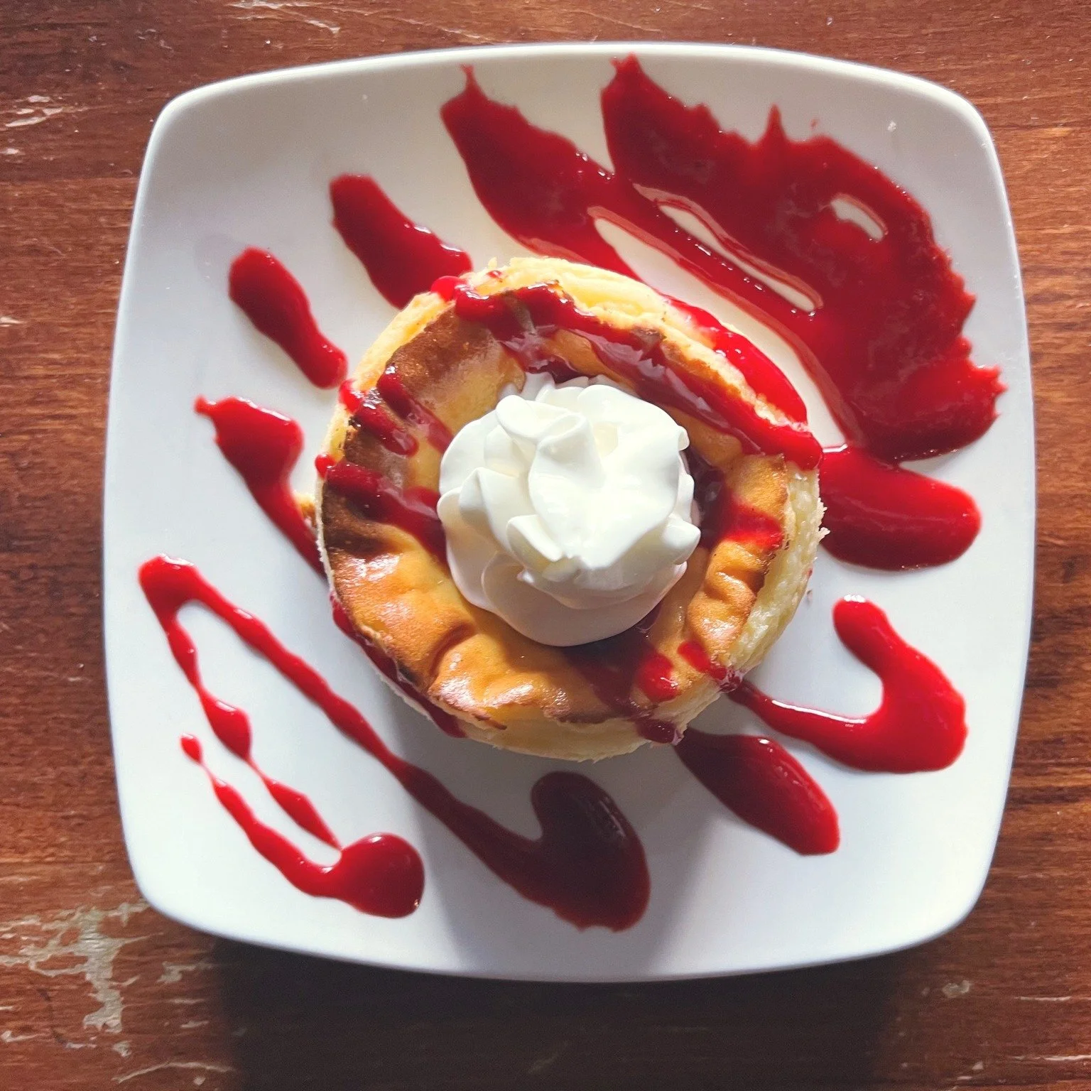 A dessert topped with whipped cream, drizzled with red fruit sauce, served on a white square plate on a wooden table.