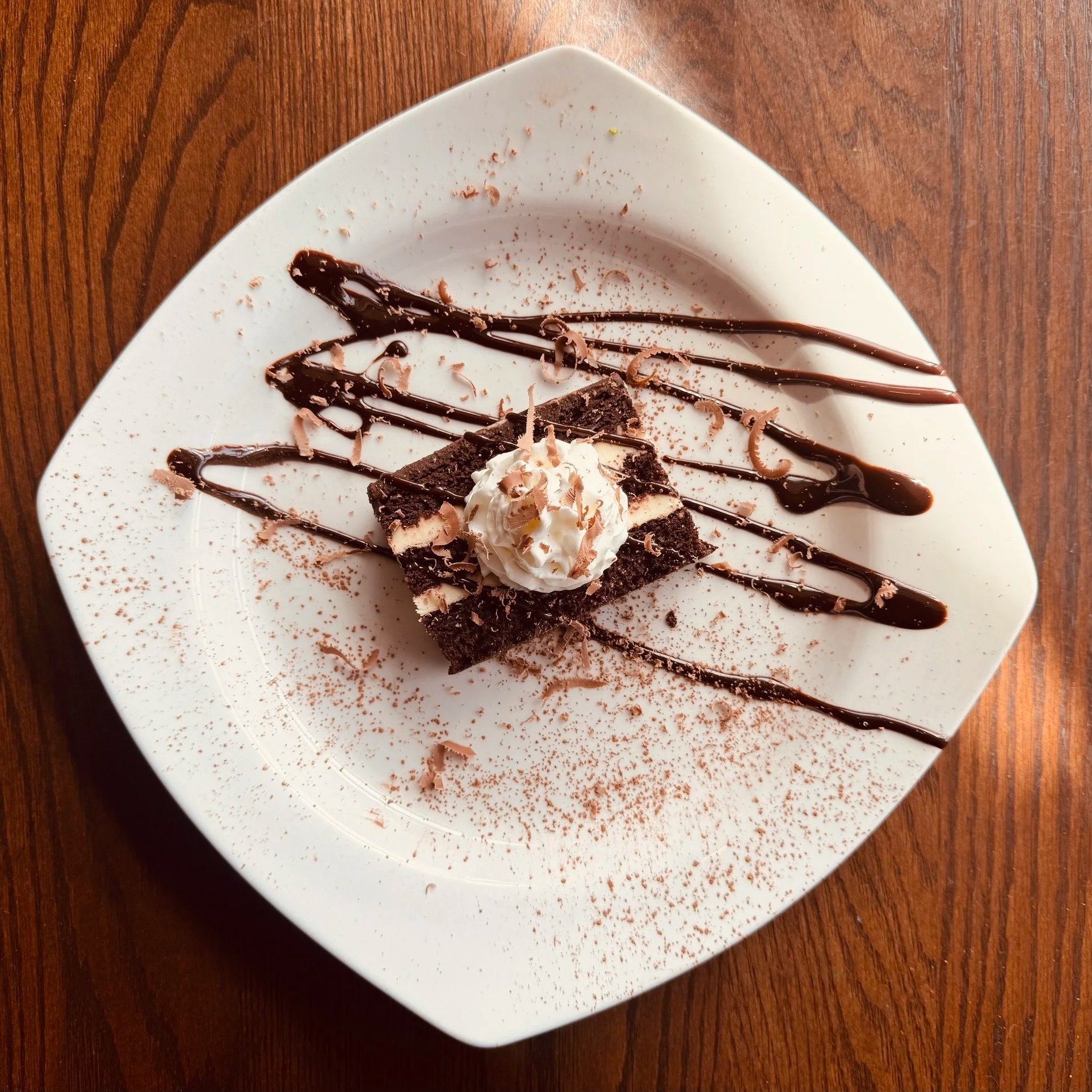 Chocolate brownies topped with whipped cream and chocolate shavings, garnished with chocolate syrup on a white square plate.