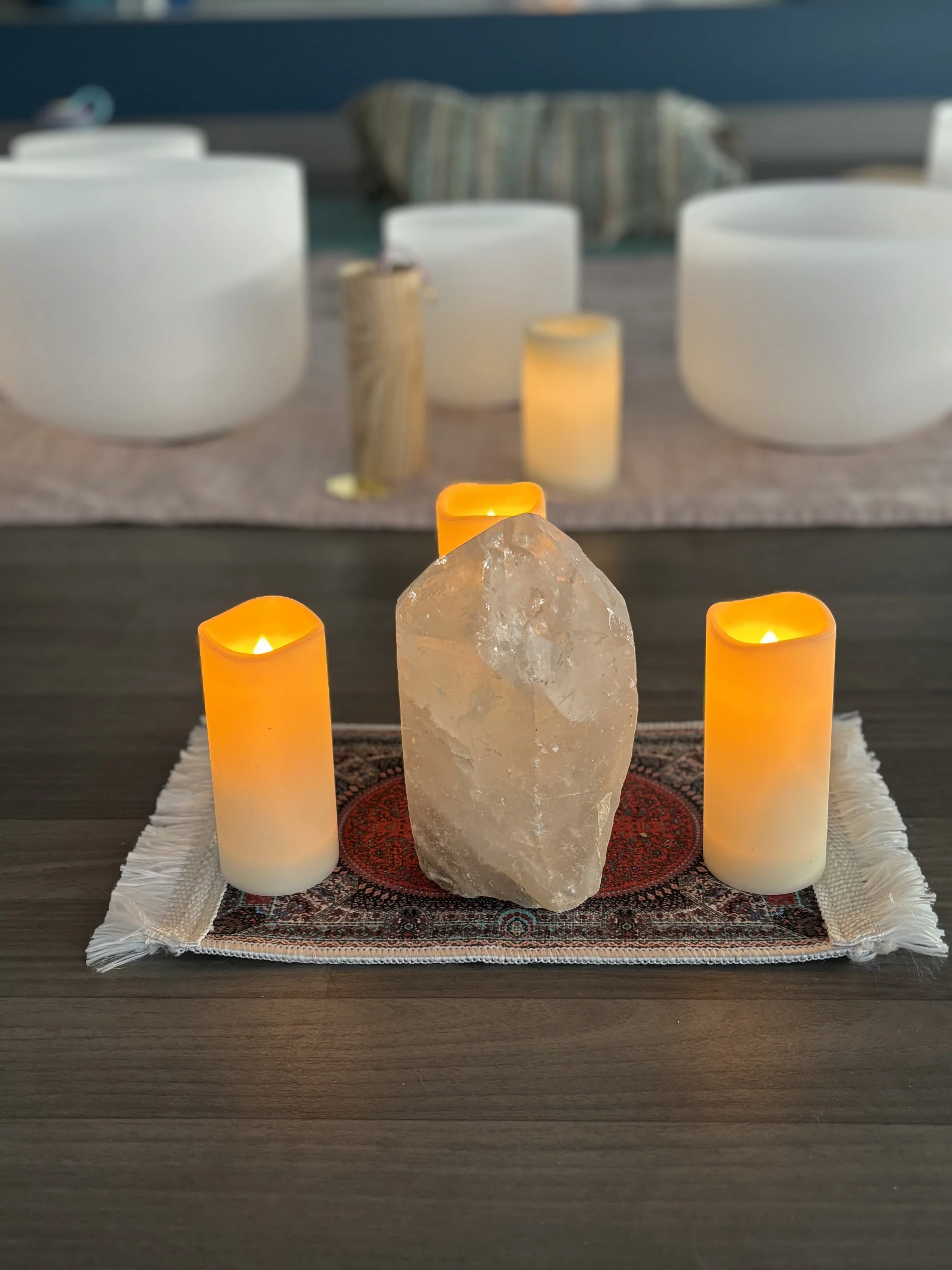A large crystal centerpiece surrounded by three orange LED candles on a decorative cloth on a table. In the background, there are four white bowls or singing bowls with candles inside, and a pillow or cushion.