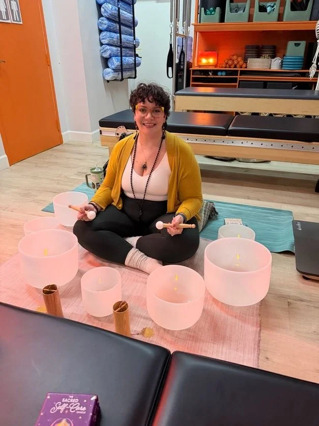 A woman sitting cross-legged on the floor in a yoga studio, surrounded by singing bowls, with a yoga mat behind her and shelves with towels and equipment in the background.