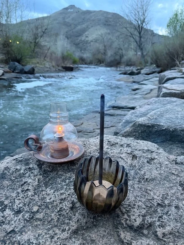 A lit oil lantern and a metallic incense holder with a stick of incense are placed on a large rock by a river, with a mountain and trees in the background.